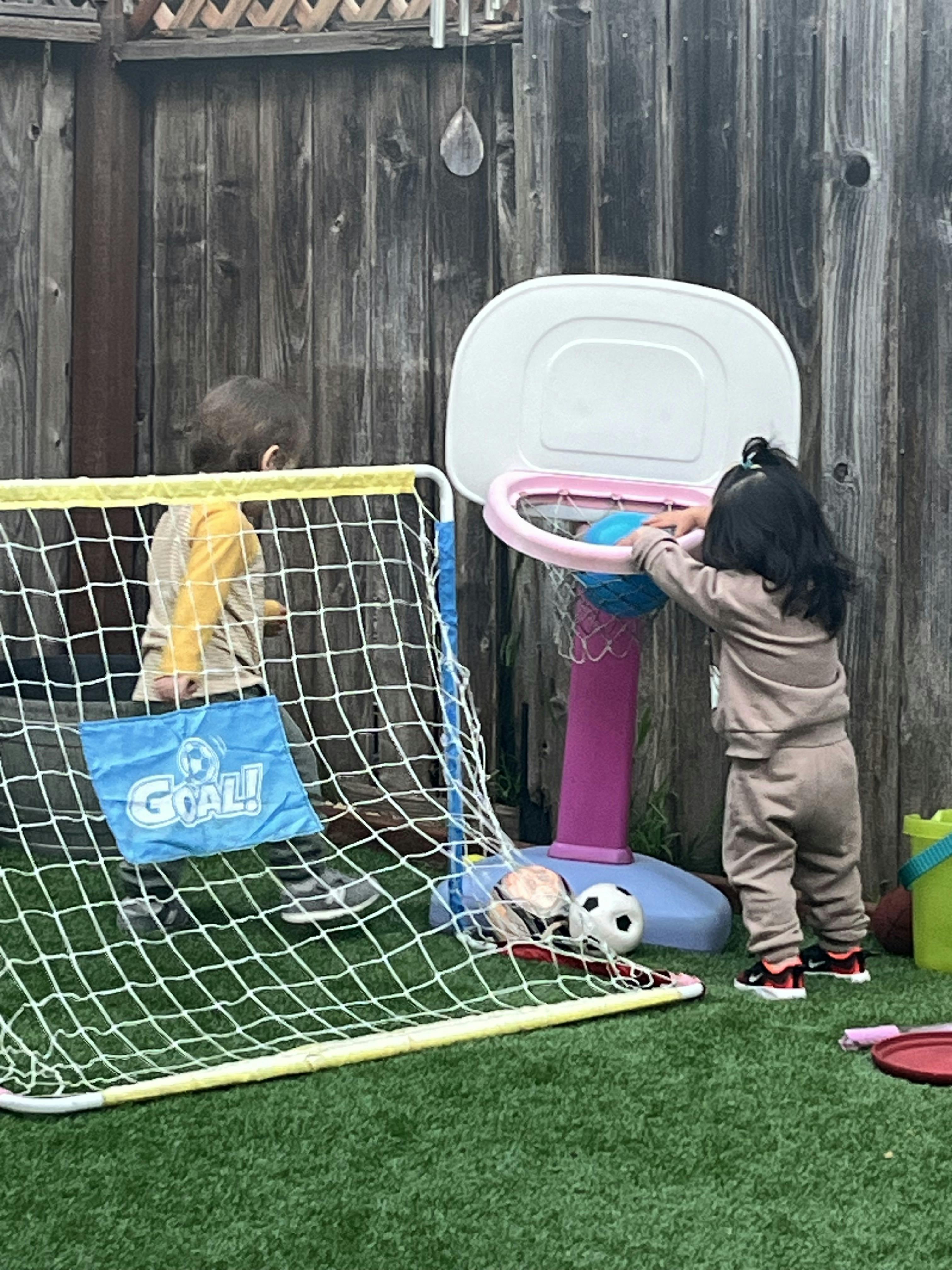 Two children playing with a soccer goal and a basketball hoop