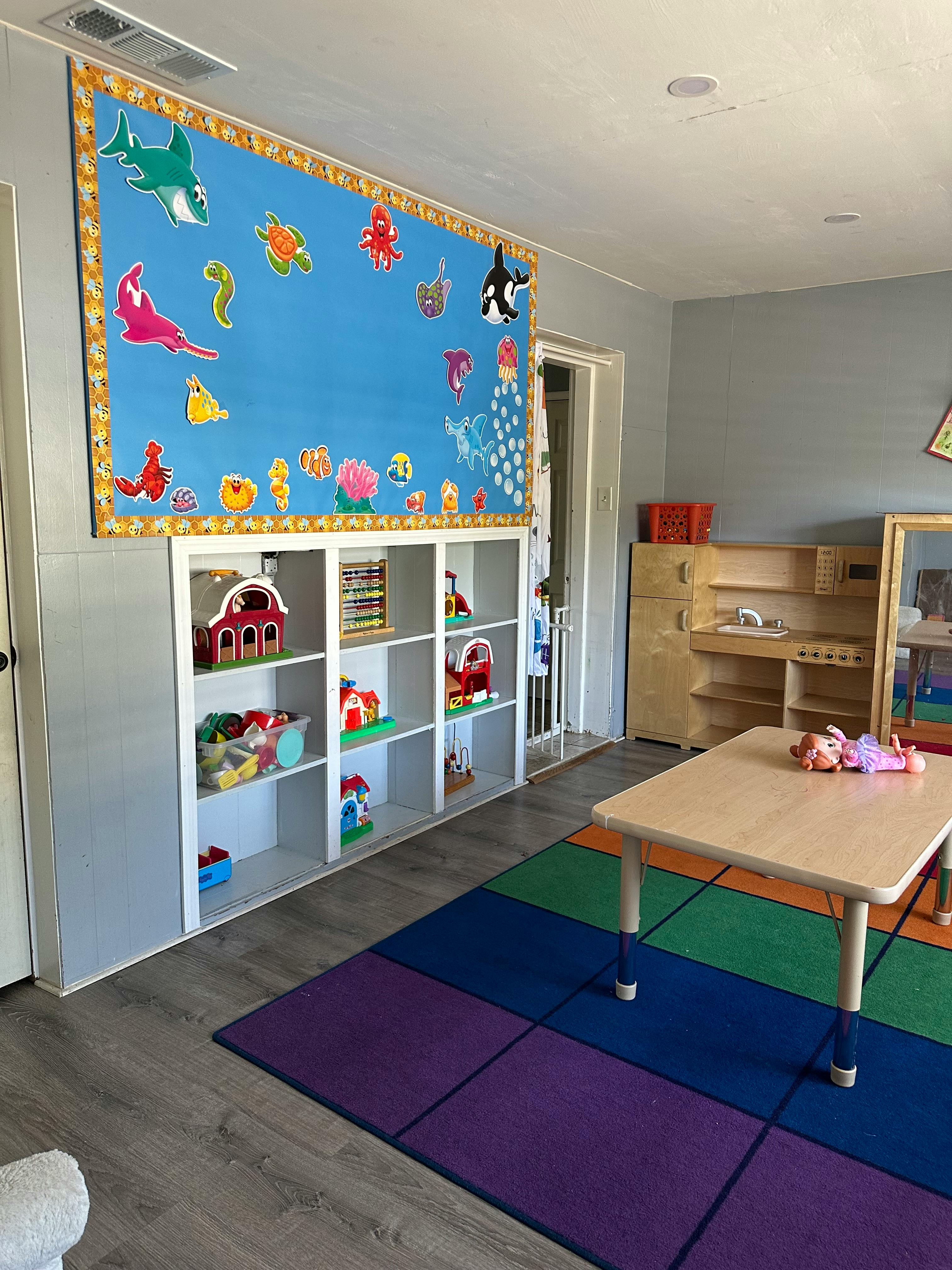 Playroom with a colorful rug, small table, shelves with toys, and a wall decoration