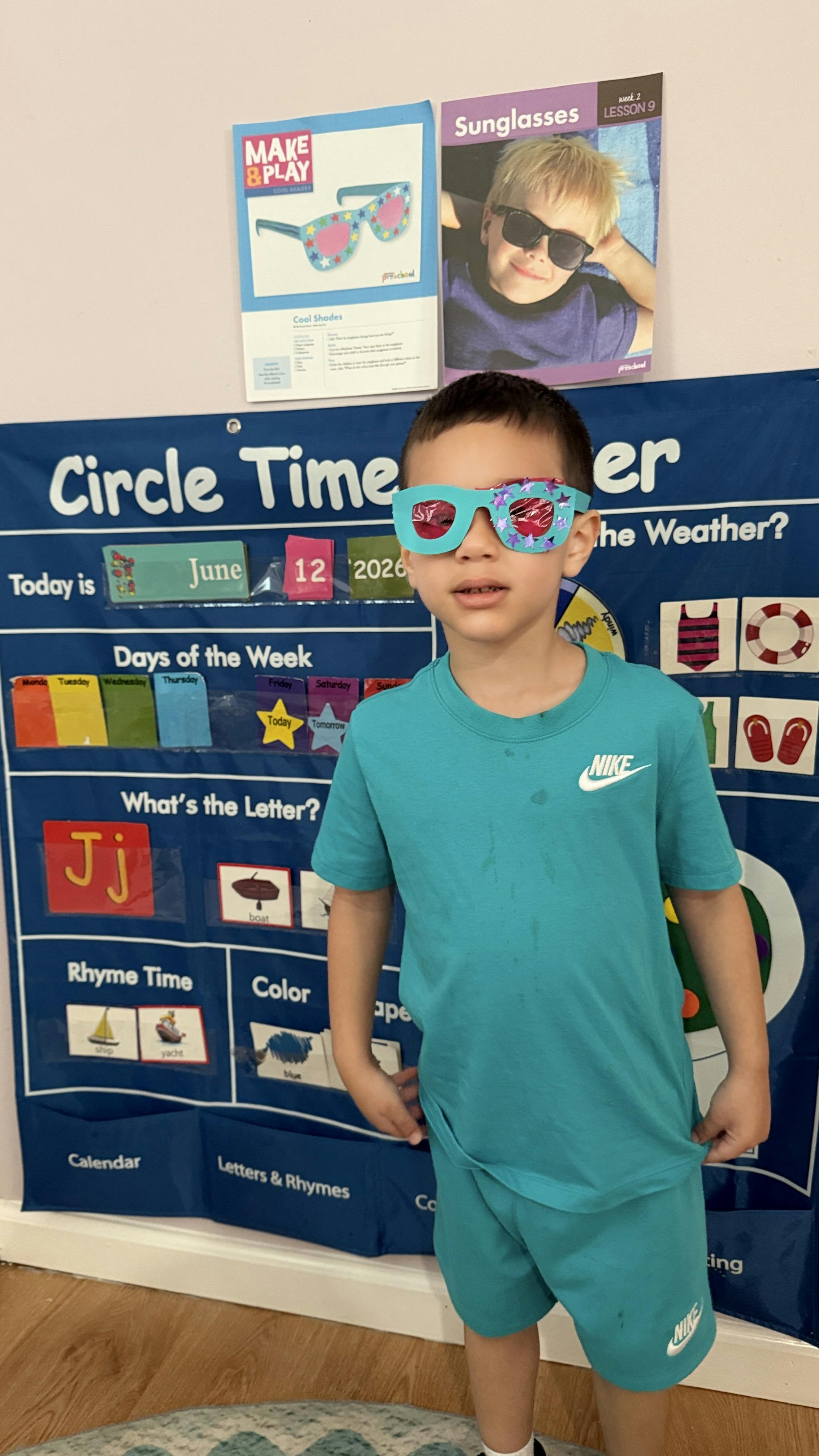 Child wearing decorated paper sunglasses in front of a classroom calendar