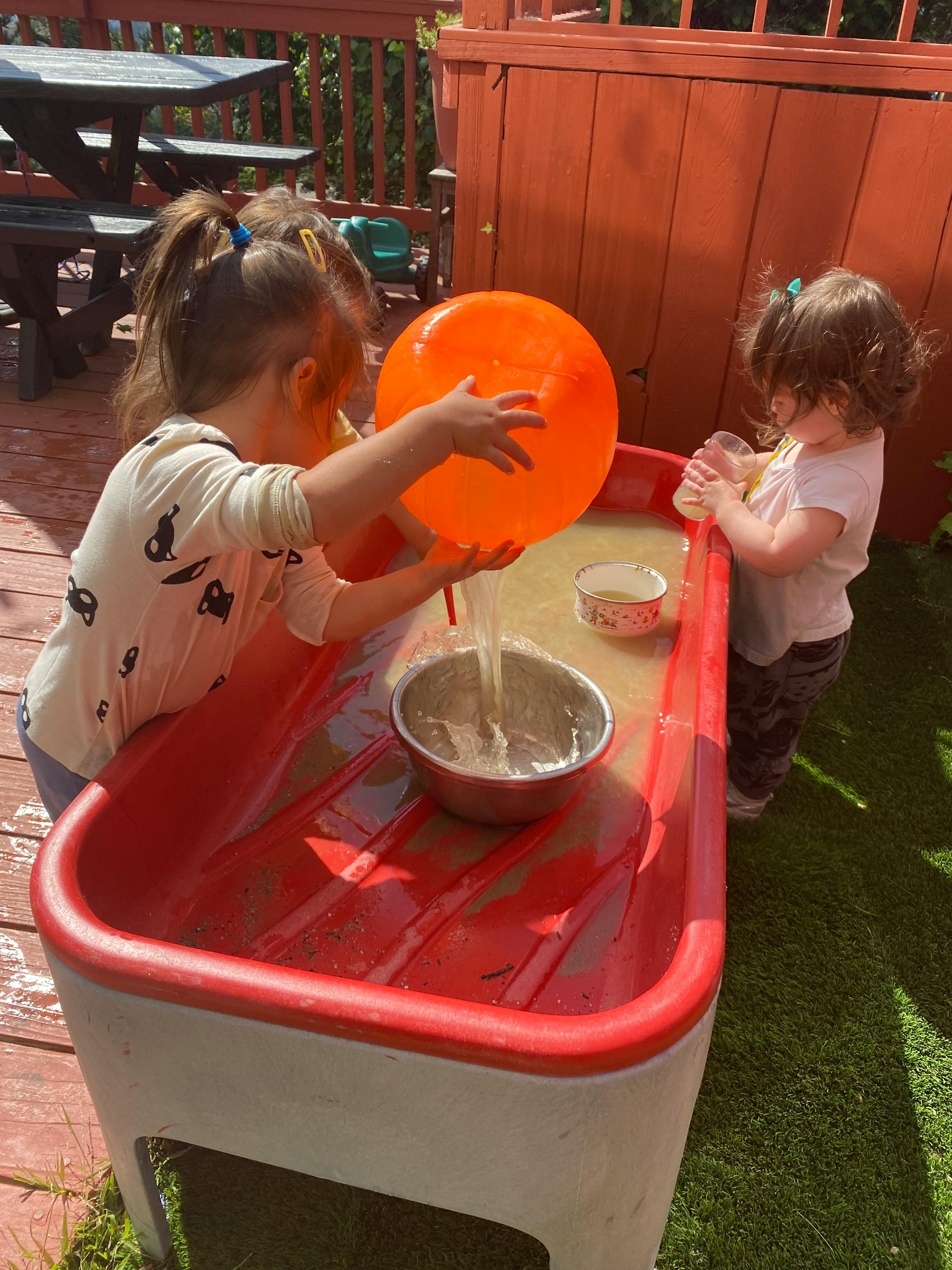 Two children playing with water in a red sensory table outdoors