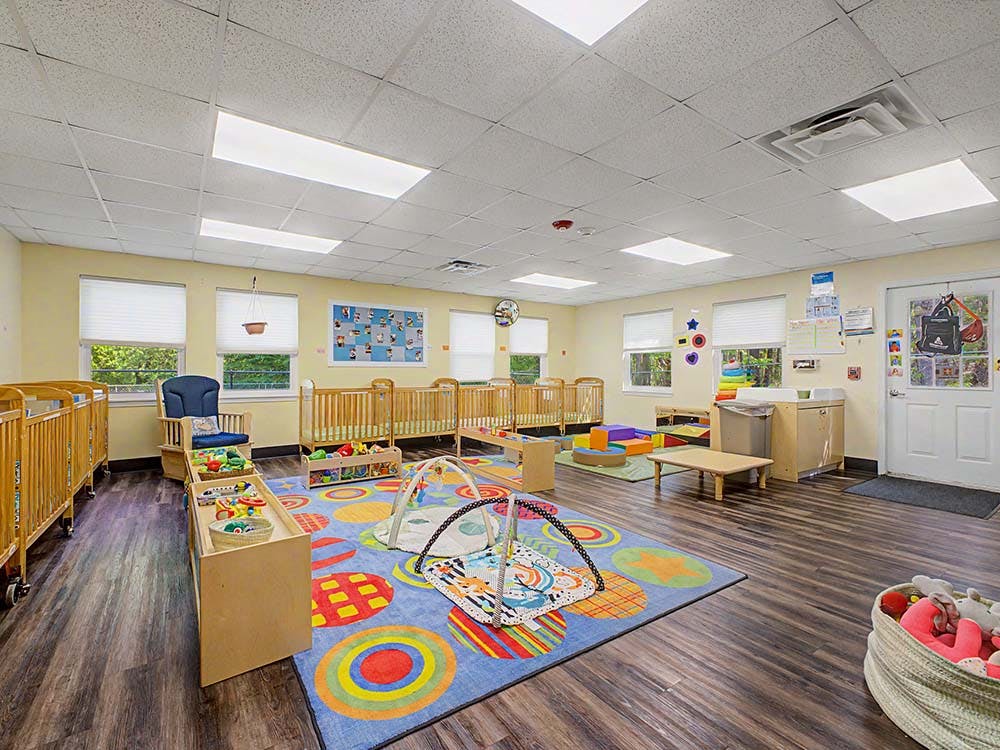 A classroom with cribs, a colorful rug, and baby play mats