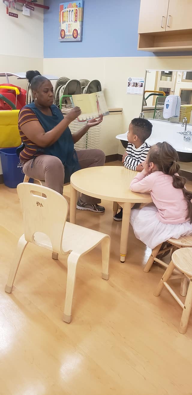 Person reading a book to two children at a small table in a classroom