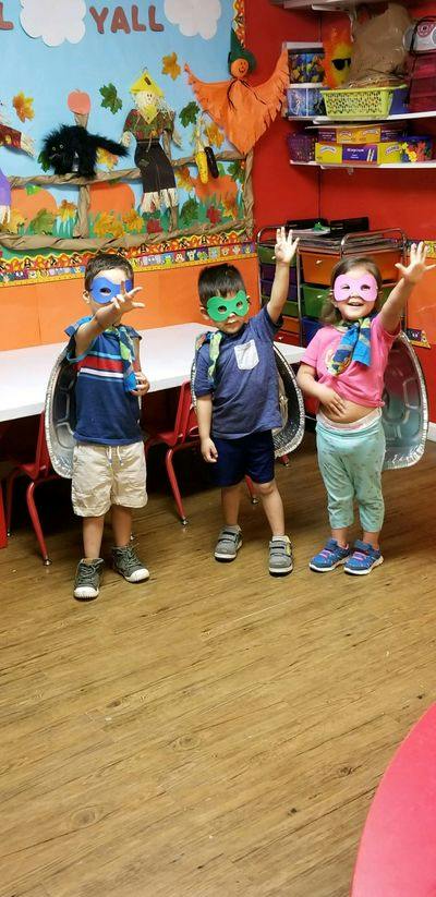 Three children wearing masks and capes, raising their hands in a classroom