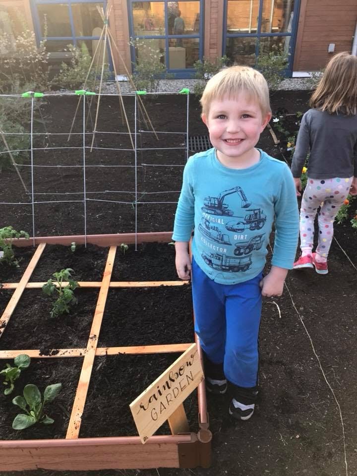 Child standing next to a garden bed with a sign that reads "Rainbow Garden"