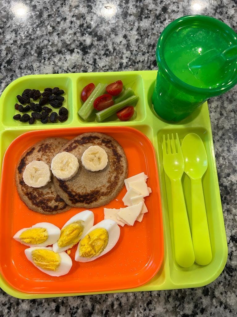 A divided plate with pancakes, hard-boiled eggs, cheese, raisins, celery, and tomatoes