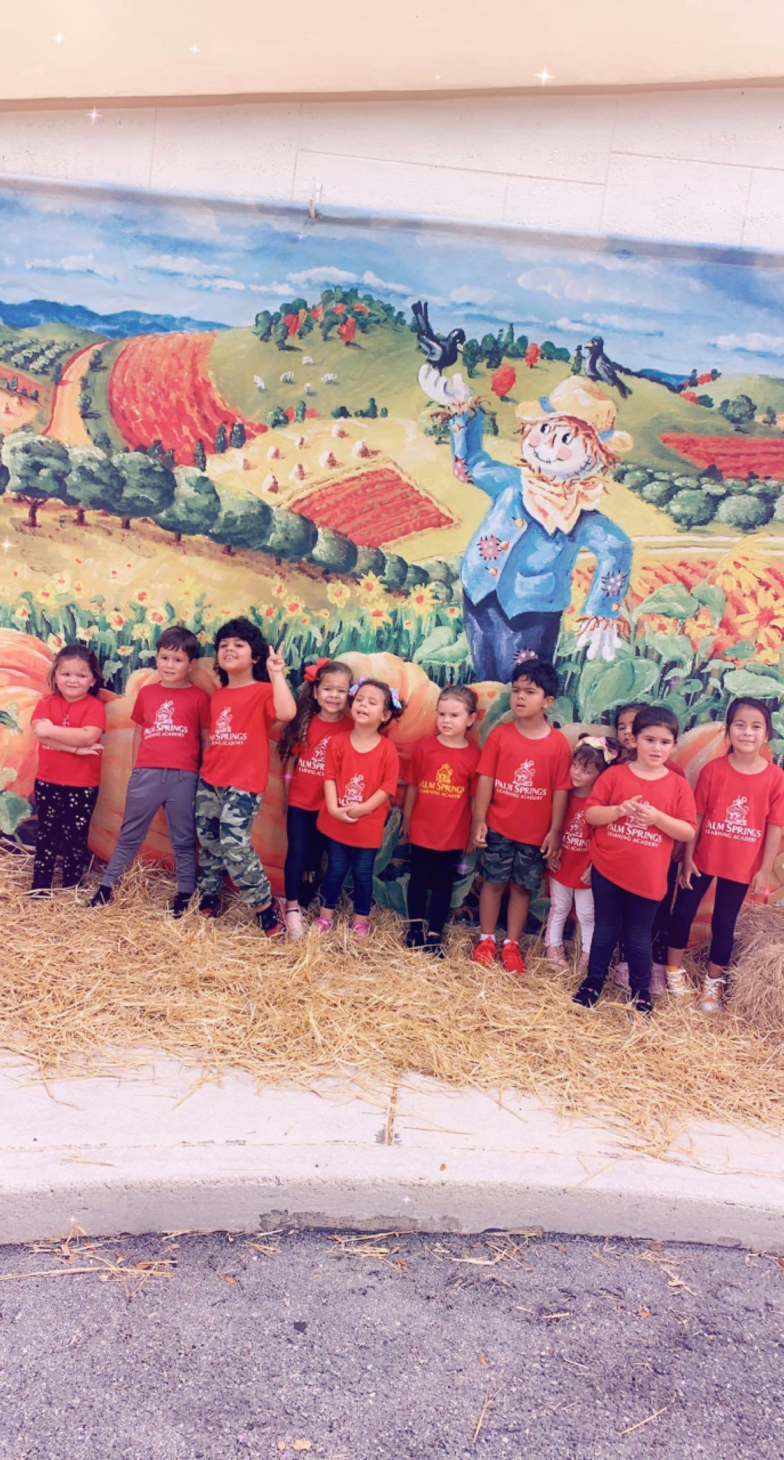 Group of children in red shirts standing in front of a mural with a scarecrow mural