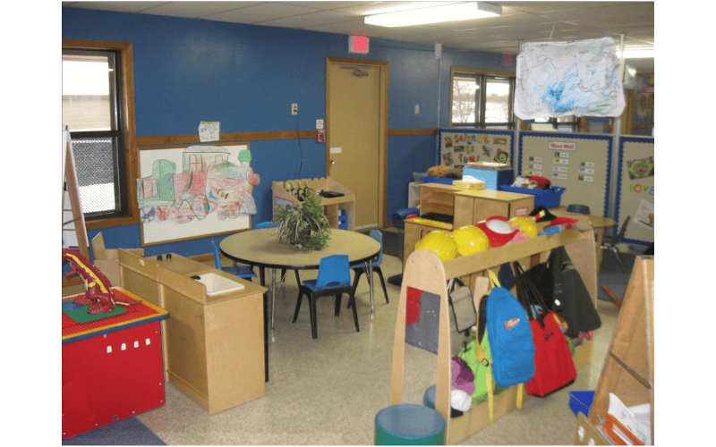 Classroom with tables, chairs, colorful drawings, and coat racks with backpacks