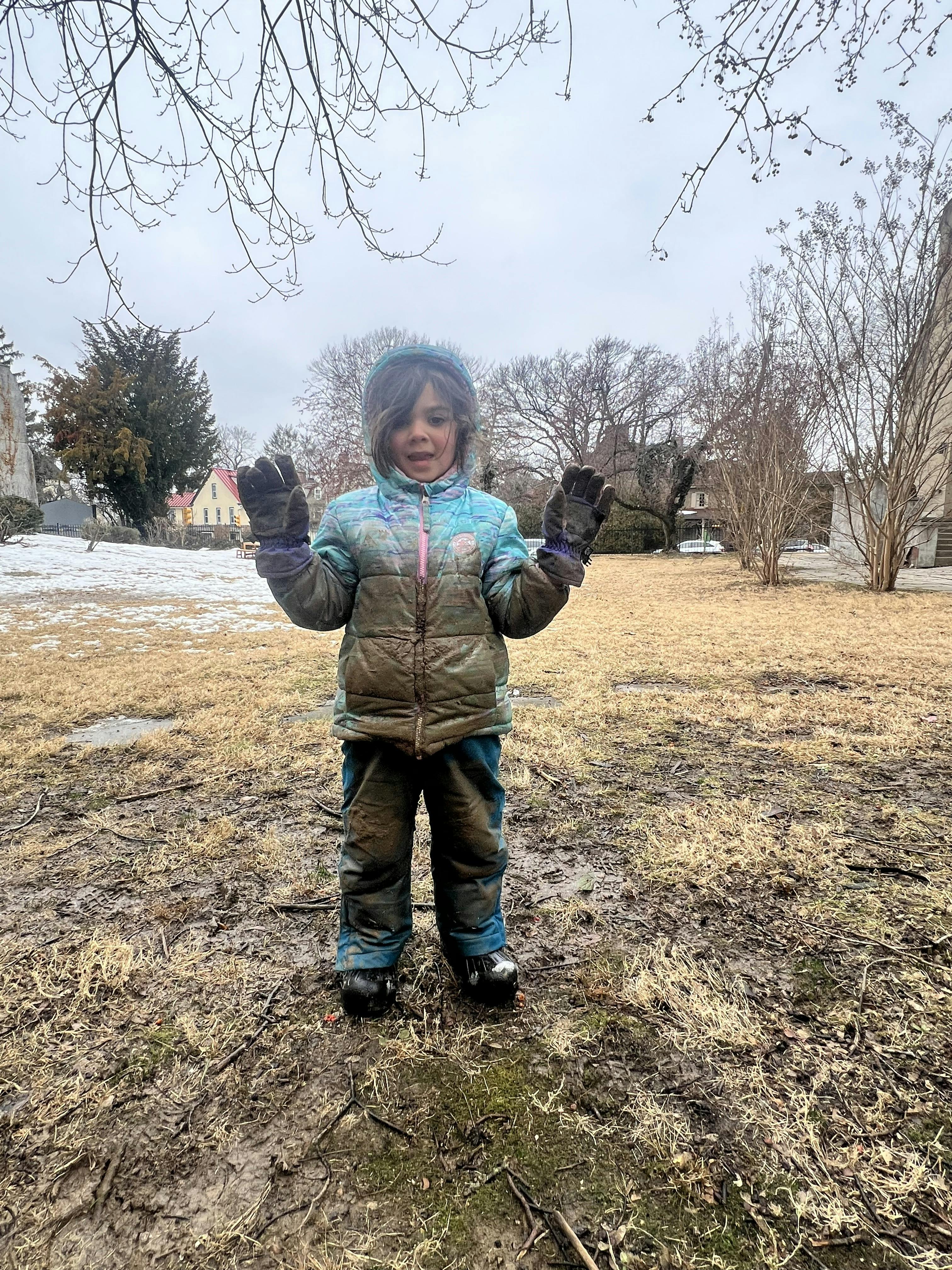 Child in a muddy winter coat and pants raising both hands, standing in a muddy field
