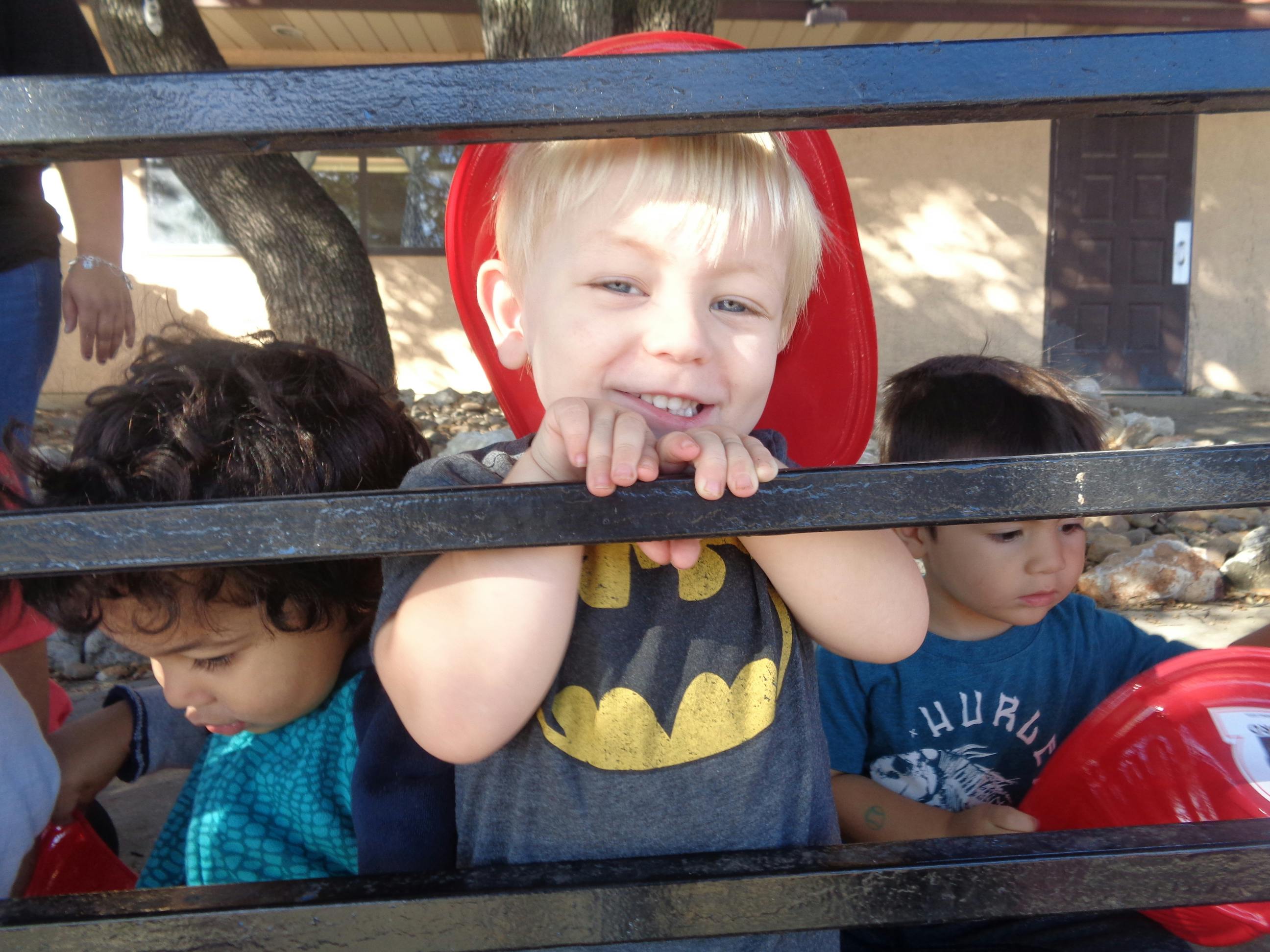 Child in red hat and Batman shirt looking through a fence with other children