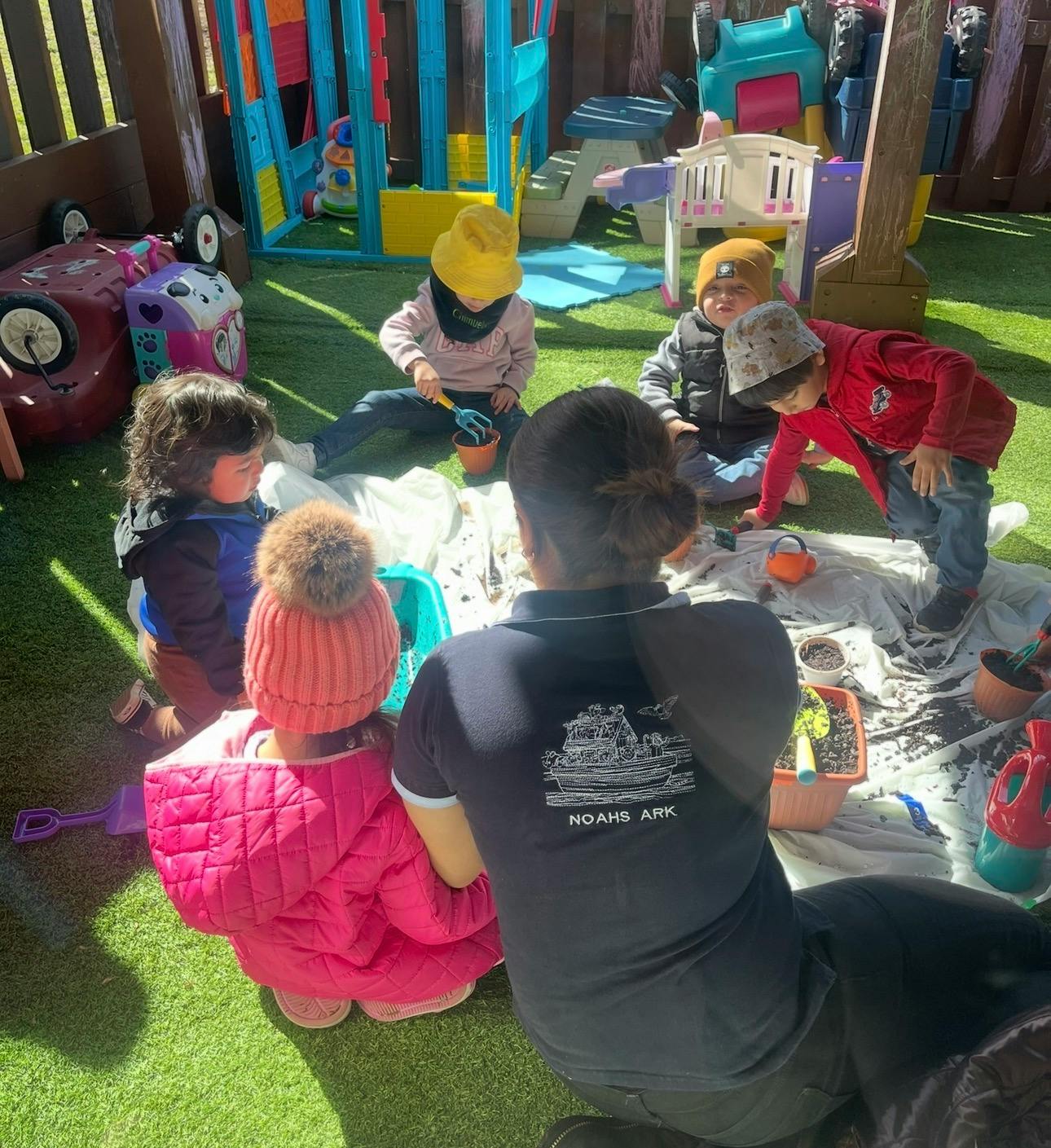 Group of children and an adult sitting on grass playing with pots and dirt