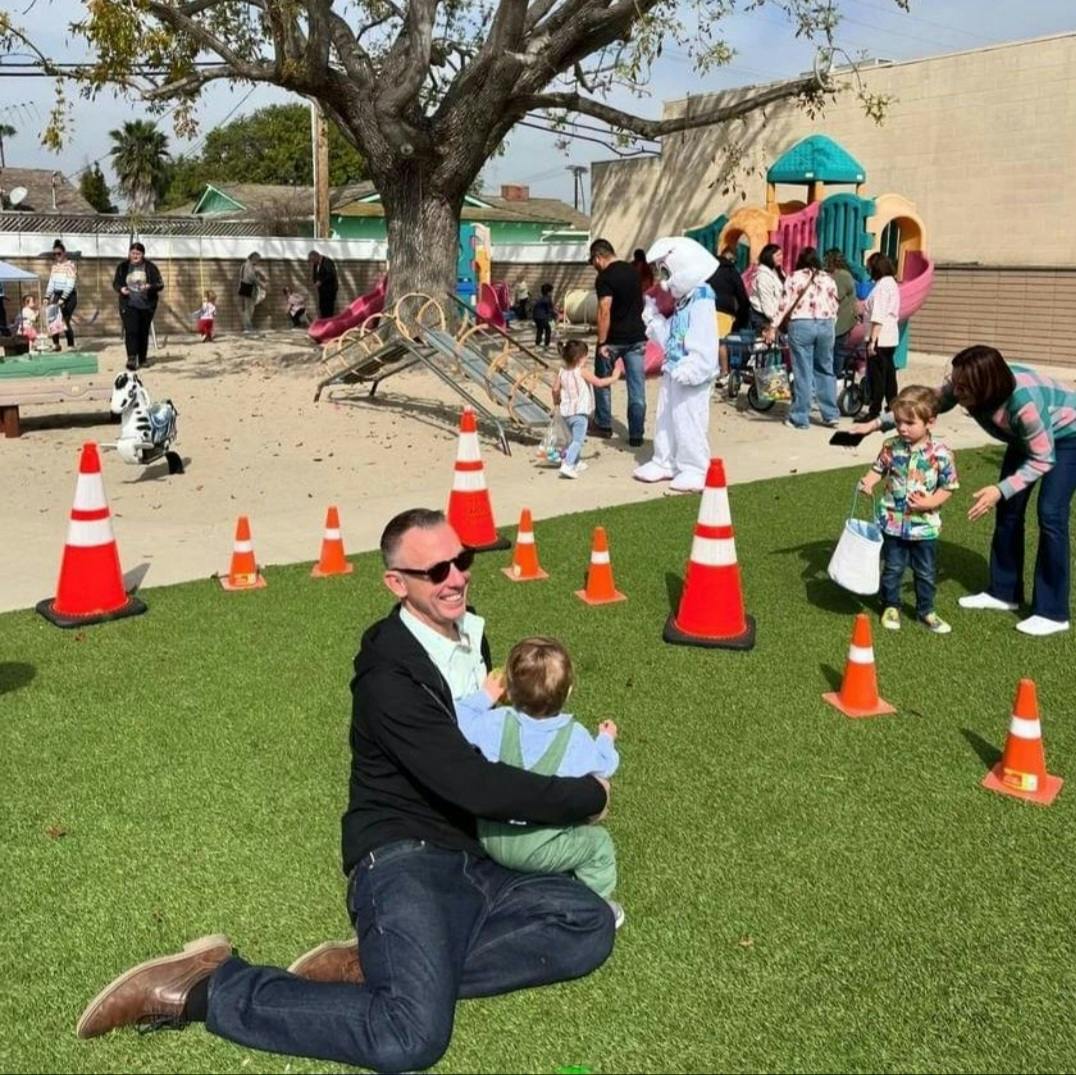 Person kneeling on grass holding a child, with other people and cones in background