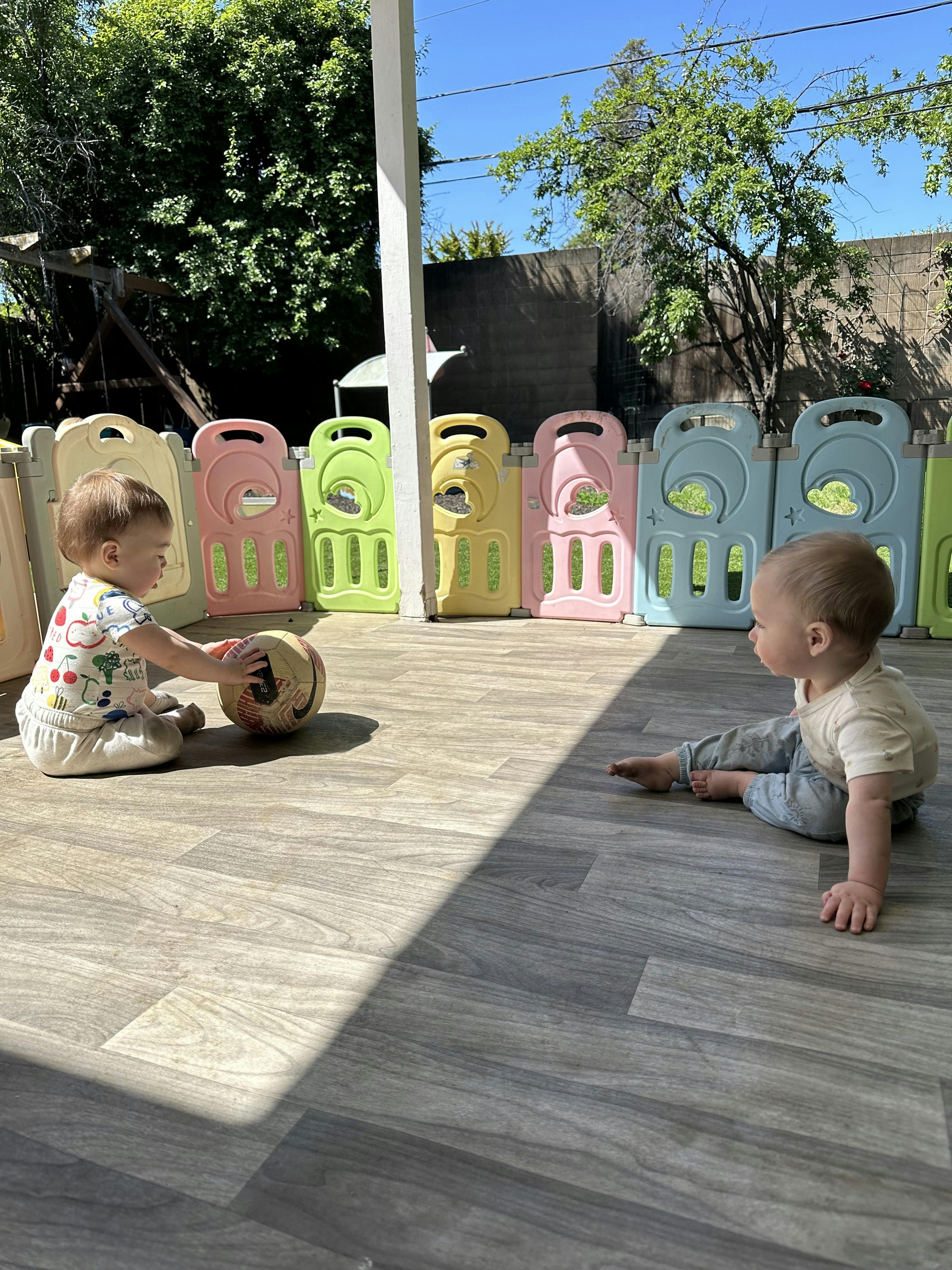 Two children on a patio, one touching a ball, the other looking on