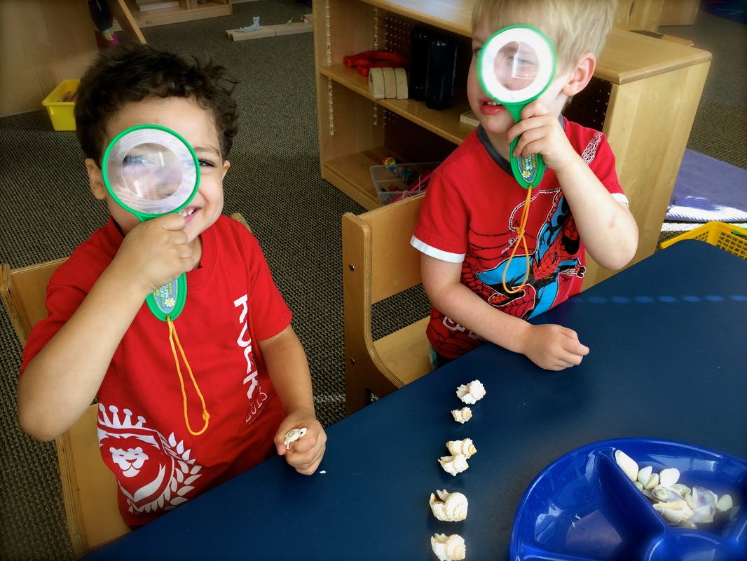 Two children looking through magnifying glasses at a blue table in a classroom