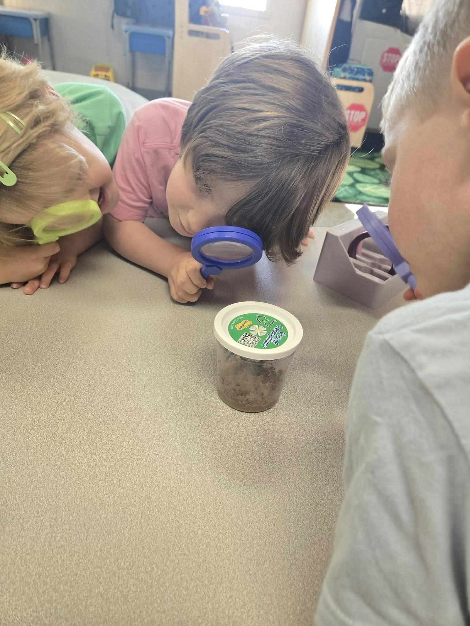 Three children looking at a container on a table, one with a magnifying glass