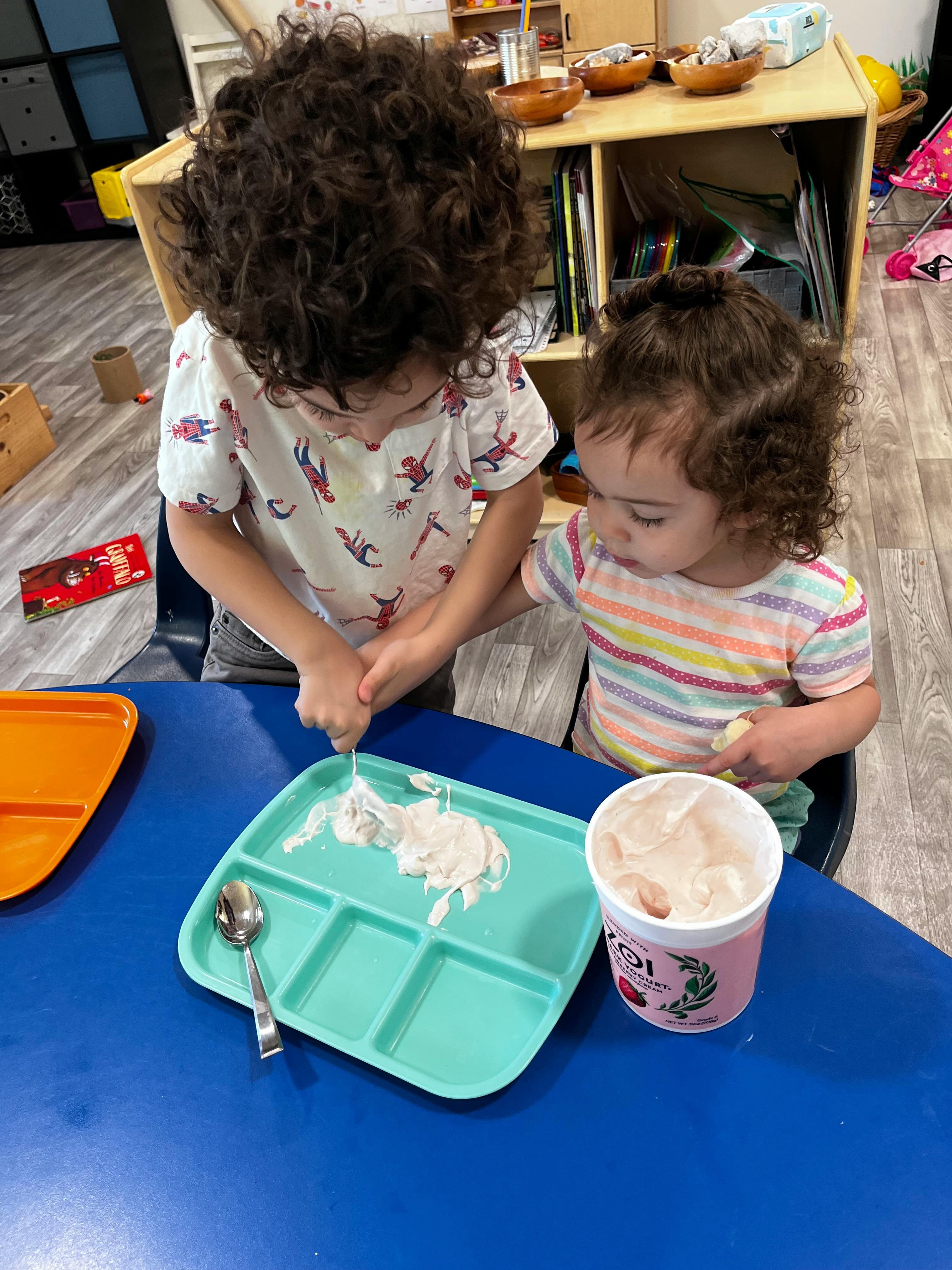 Two children at a table, one scooping from a container onto a tray