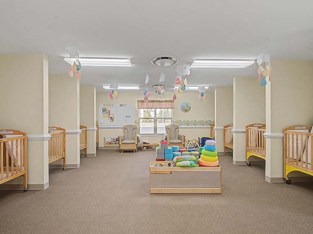 Cribs lining the walls of a daycare or nursery room with toys in the center