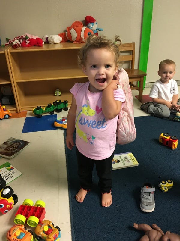 Child standing in a room with toys, another child sitting in the background