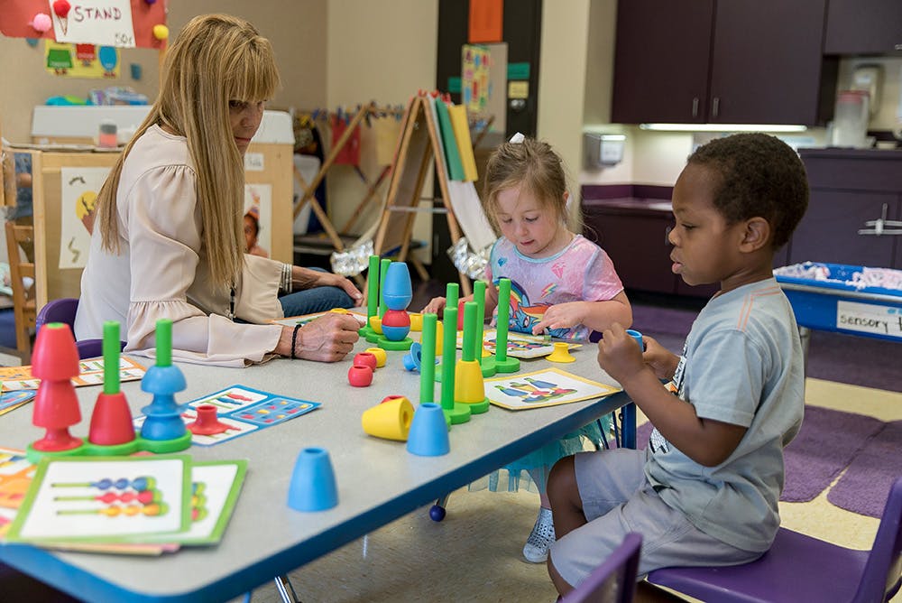 Adult and two children playing with colorful blocks at a table in a classroom