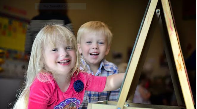 Two children smiling, one reaching towards an easel