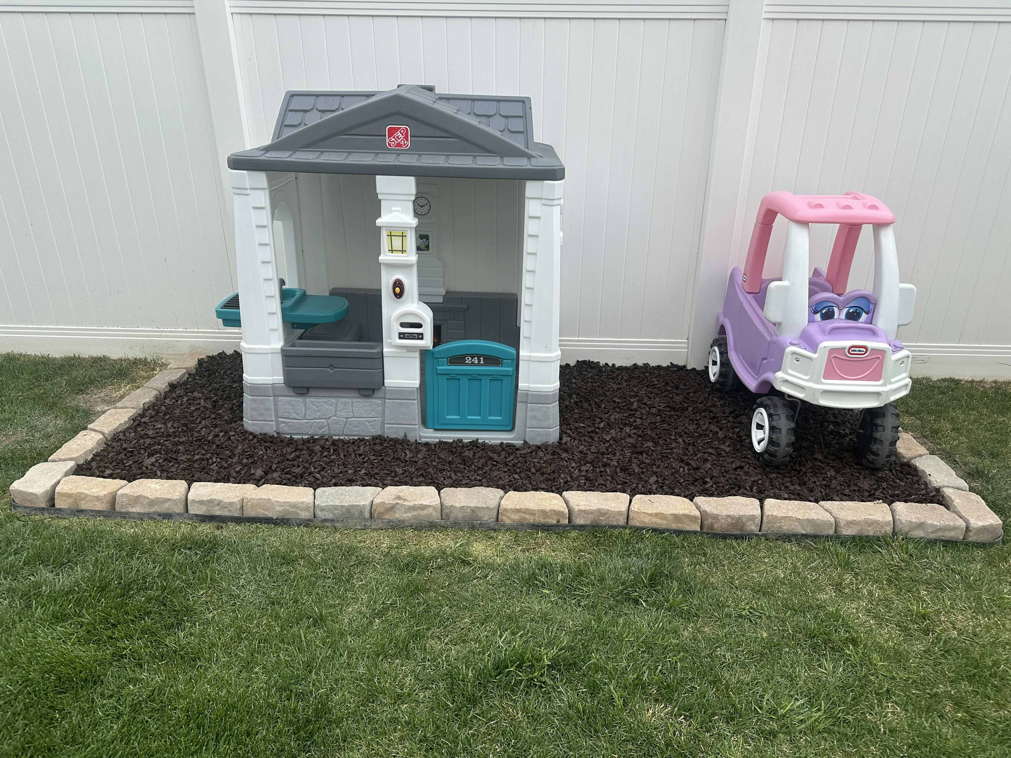 Plastic playhouse and toy car on a bed of mulch with a stone border