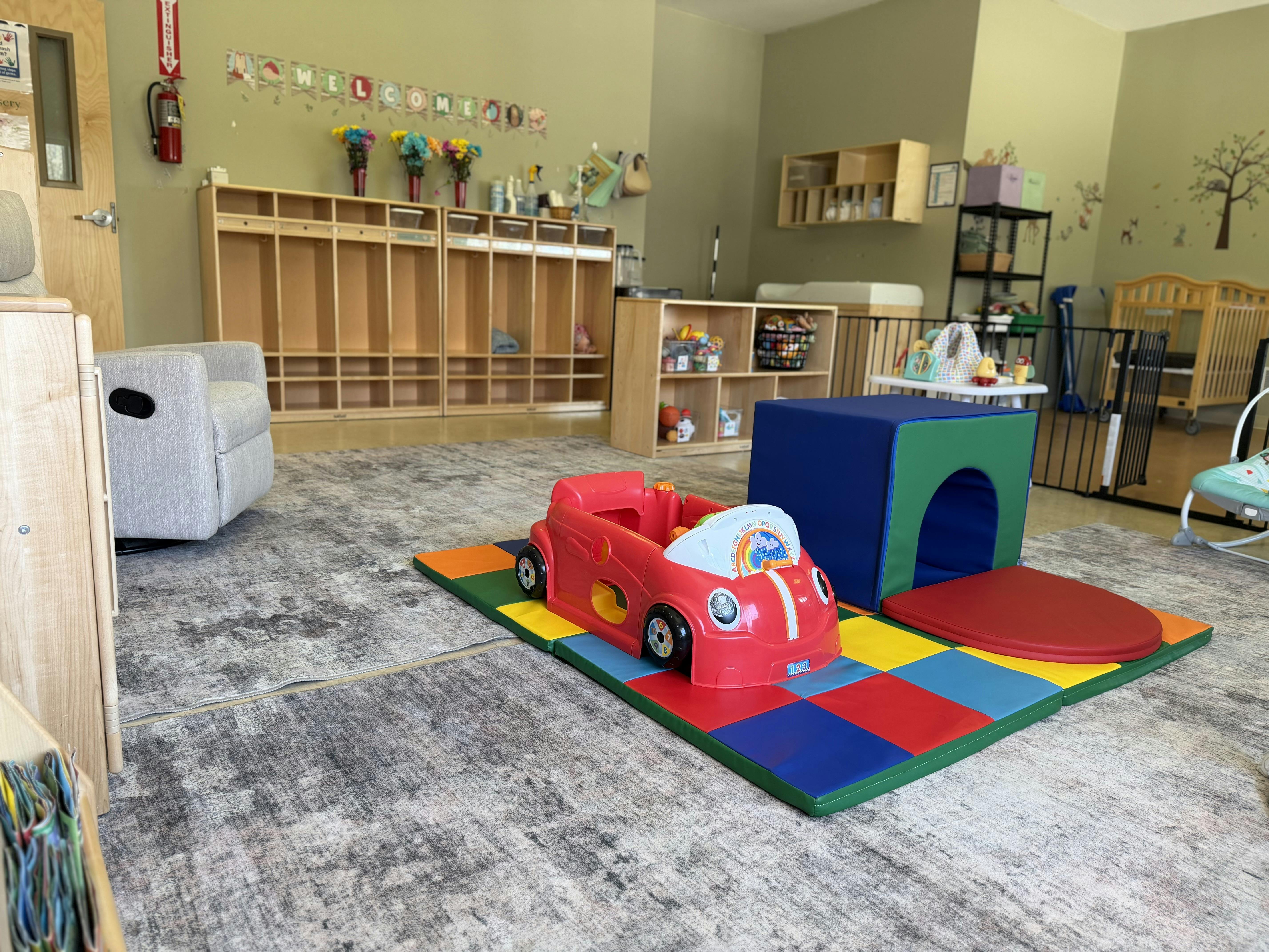 Playroom with a red toy car on colorful floor mats, cubbies, and cribs