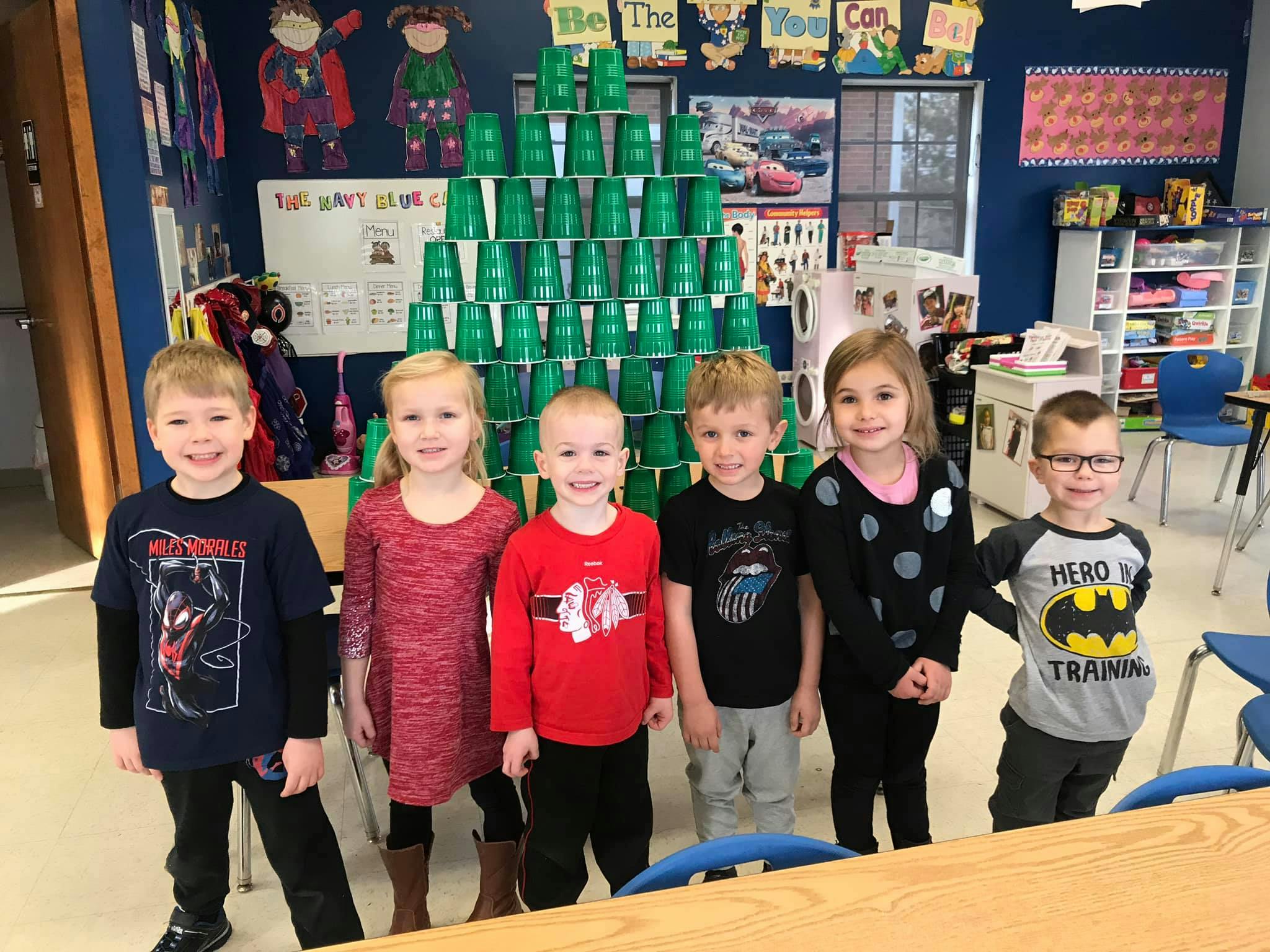 Six children standing in a classroom with a large stack of green cups behind them