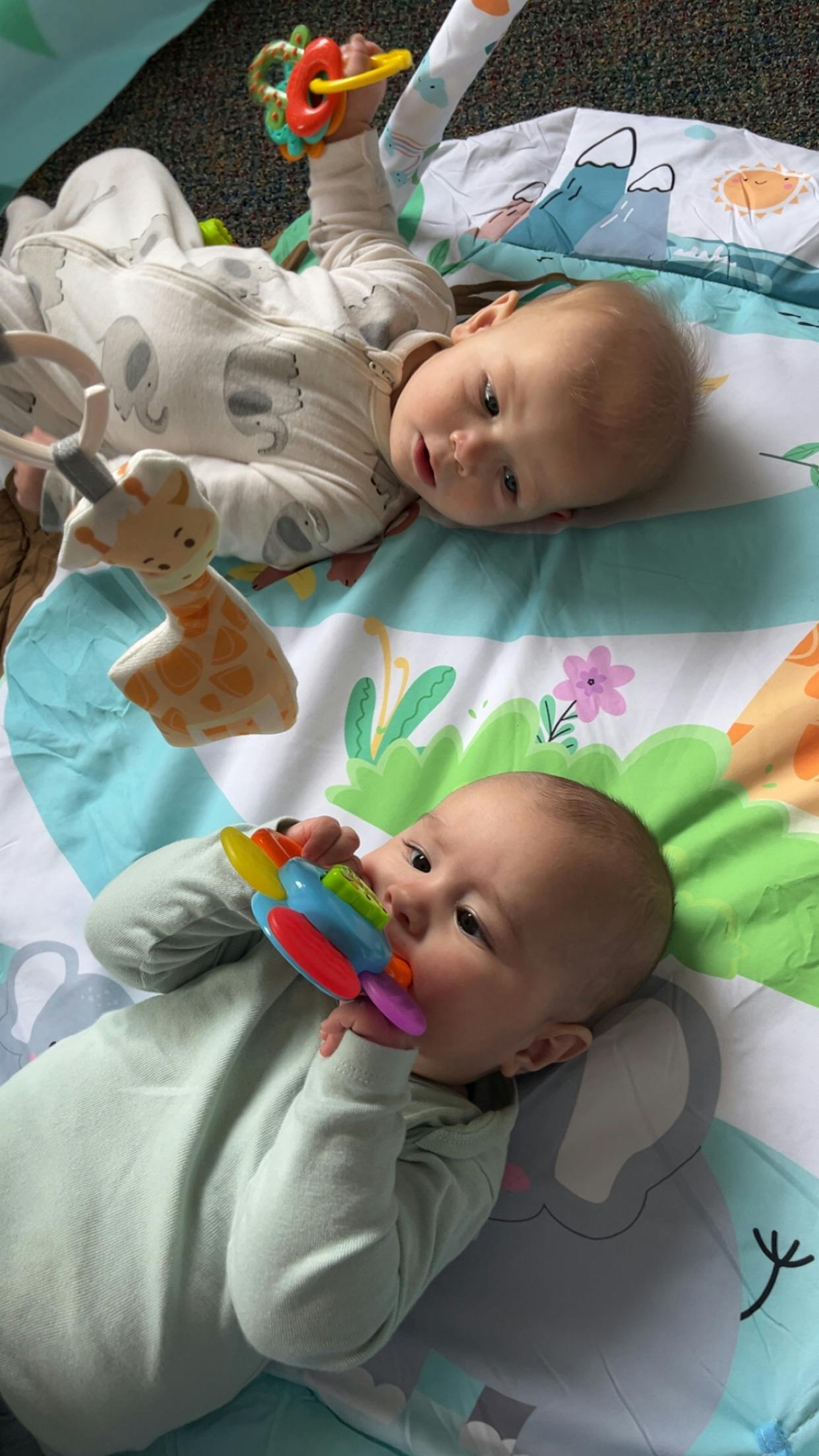 Two infants lying on a playmat, one chewing a colorful toy