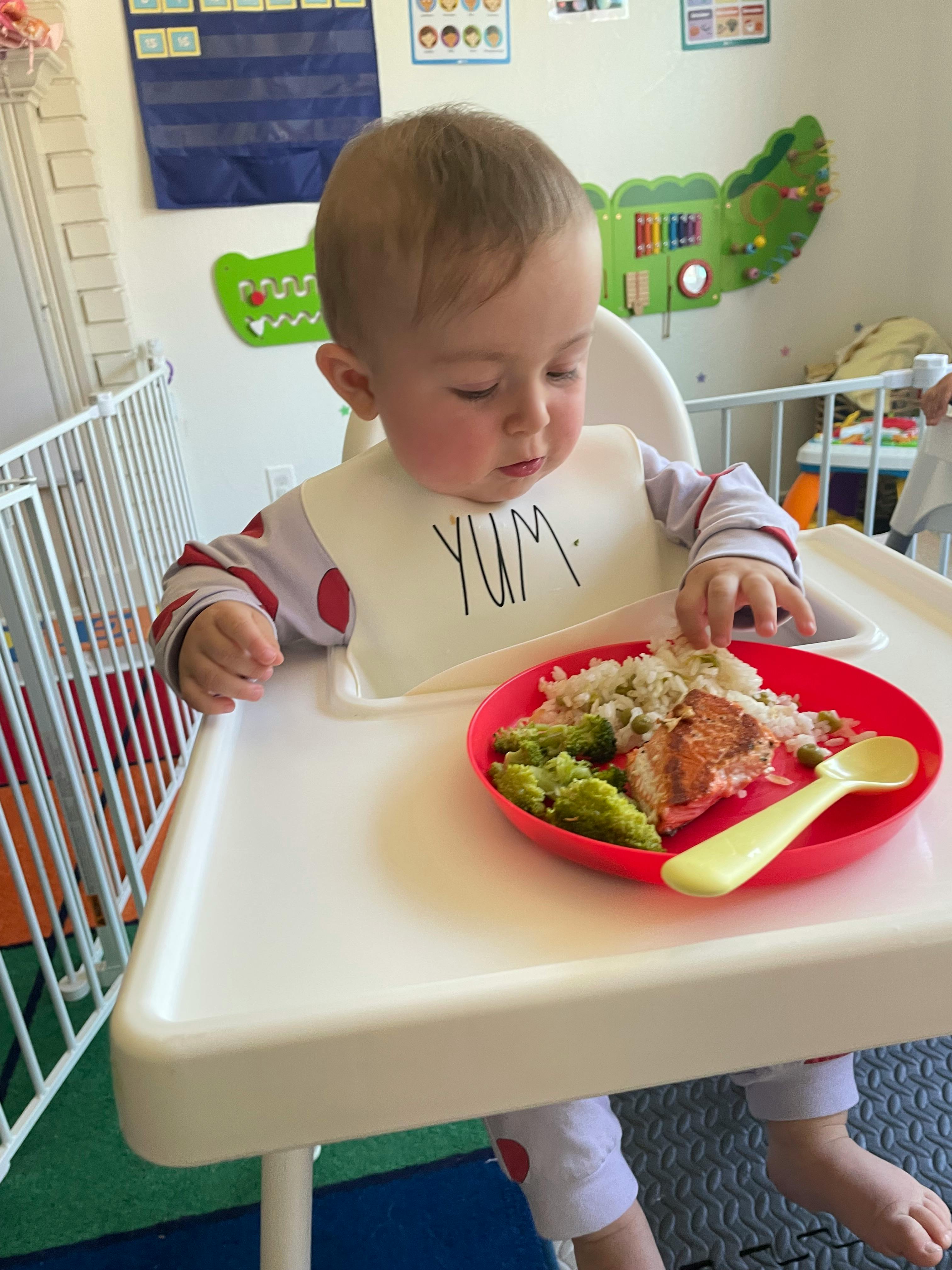 Child in high chair with bib that says YUM, looking at a plate of food