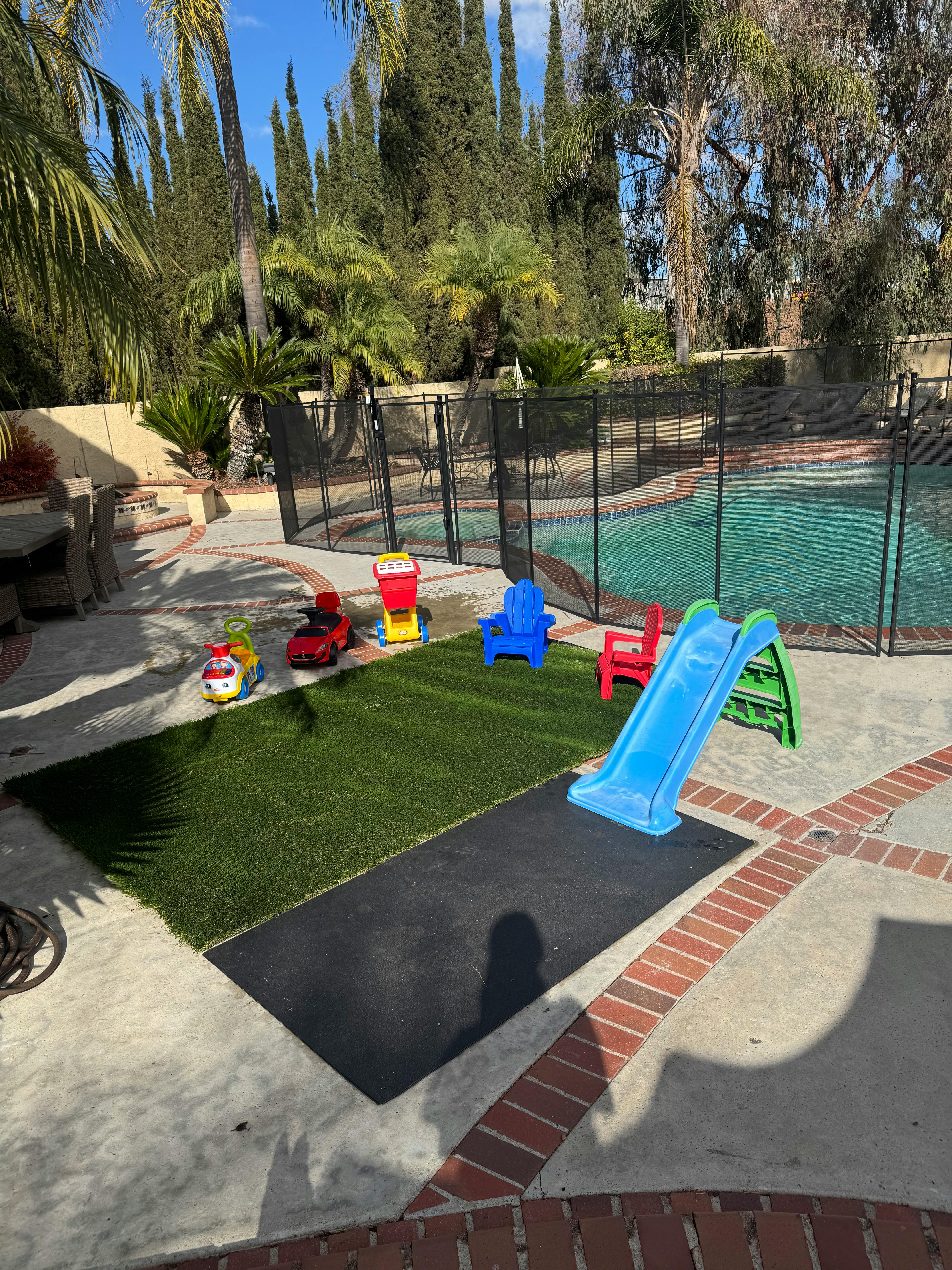 Play area with artificial grass, toys, and a slide next to a fenced pool