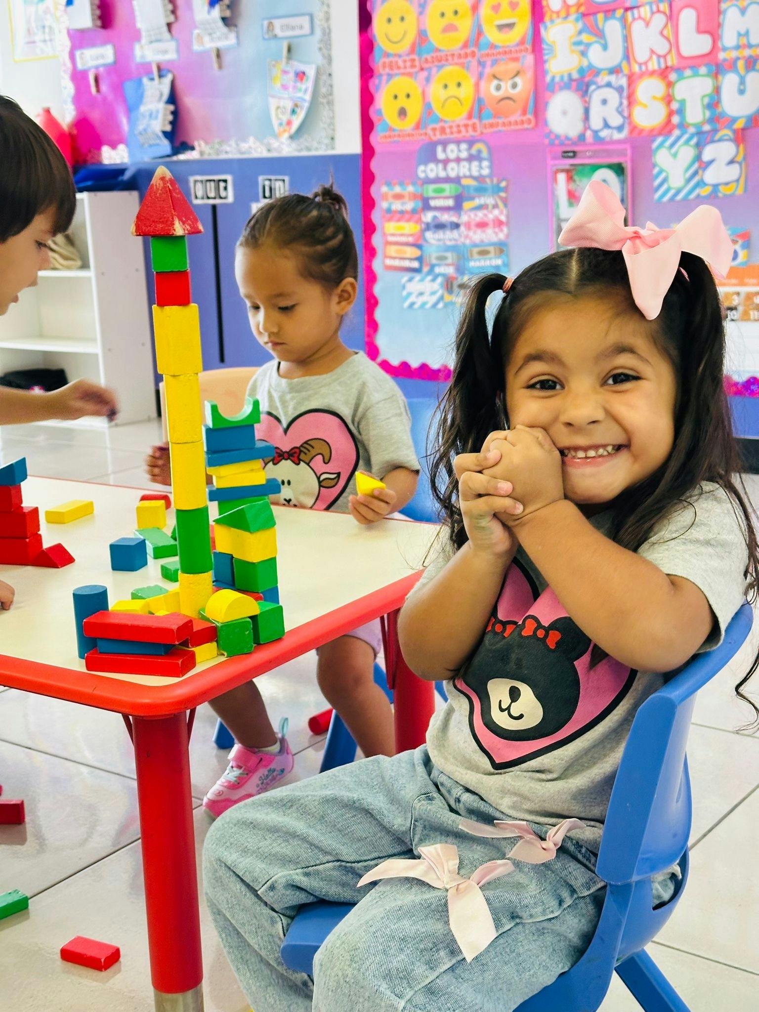 Child playing with blocks at a table, another child smiling at the camera