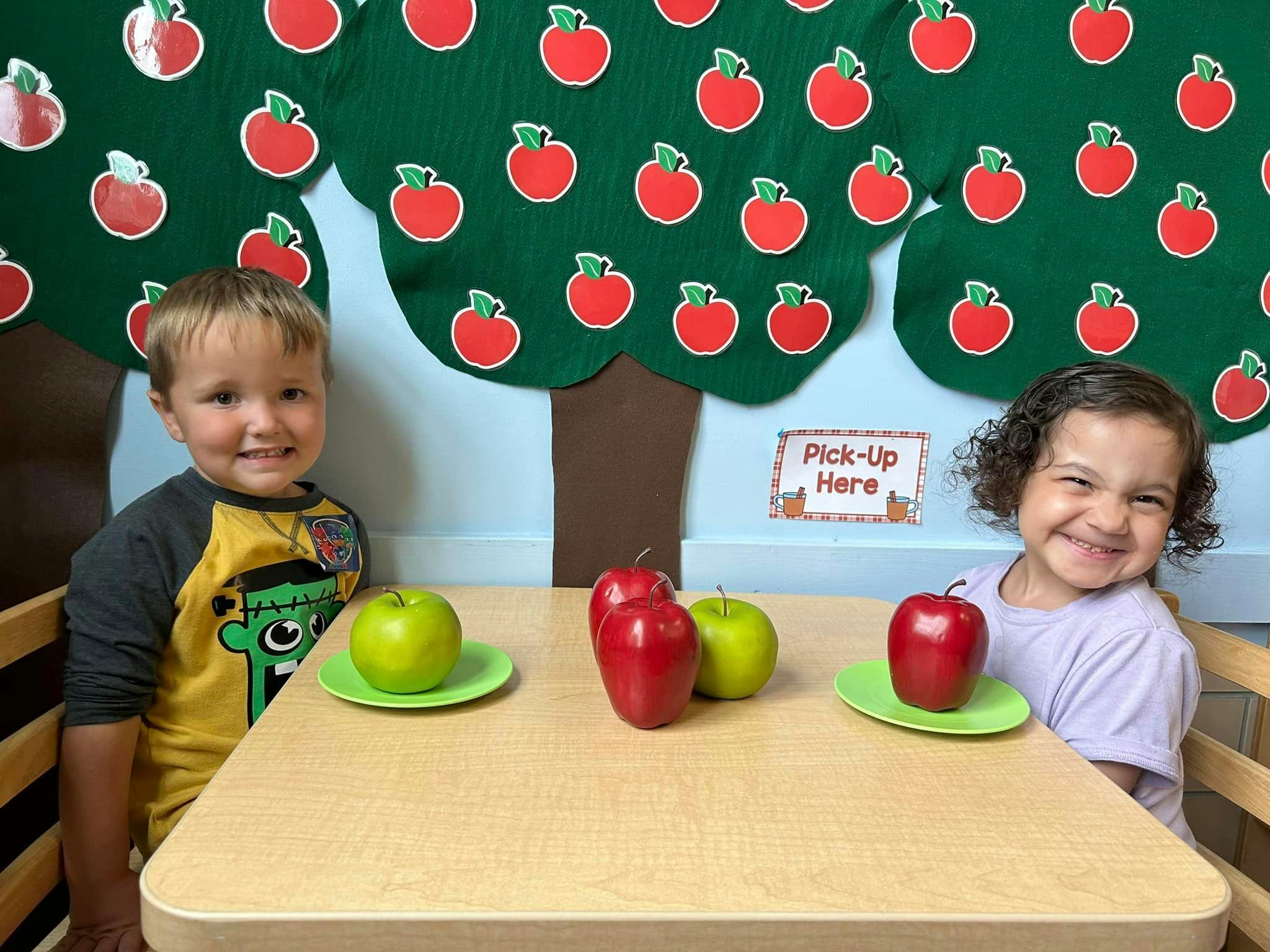 Two children sitting at a table with apples, apple tree decoration on wall