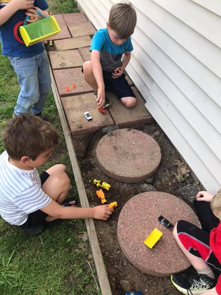 Three children playing with toy cars on a brick path next to a house