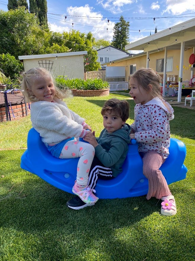 Three children playing on a blue plastic seesaw in a backyard