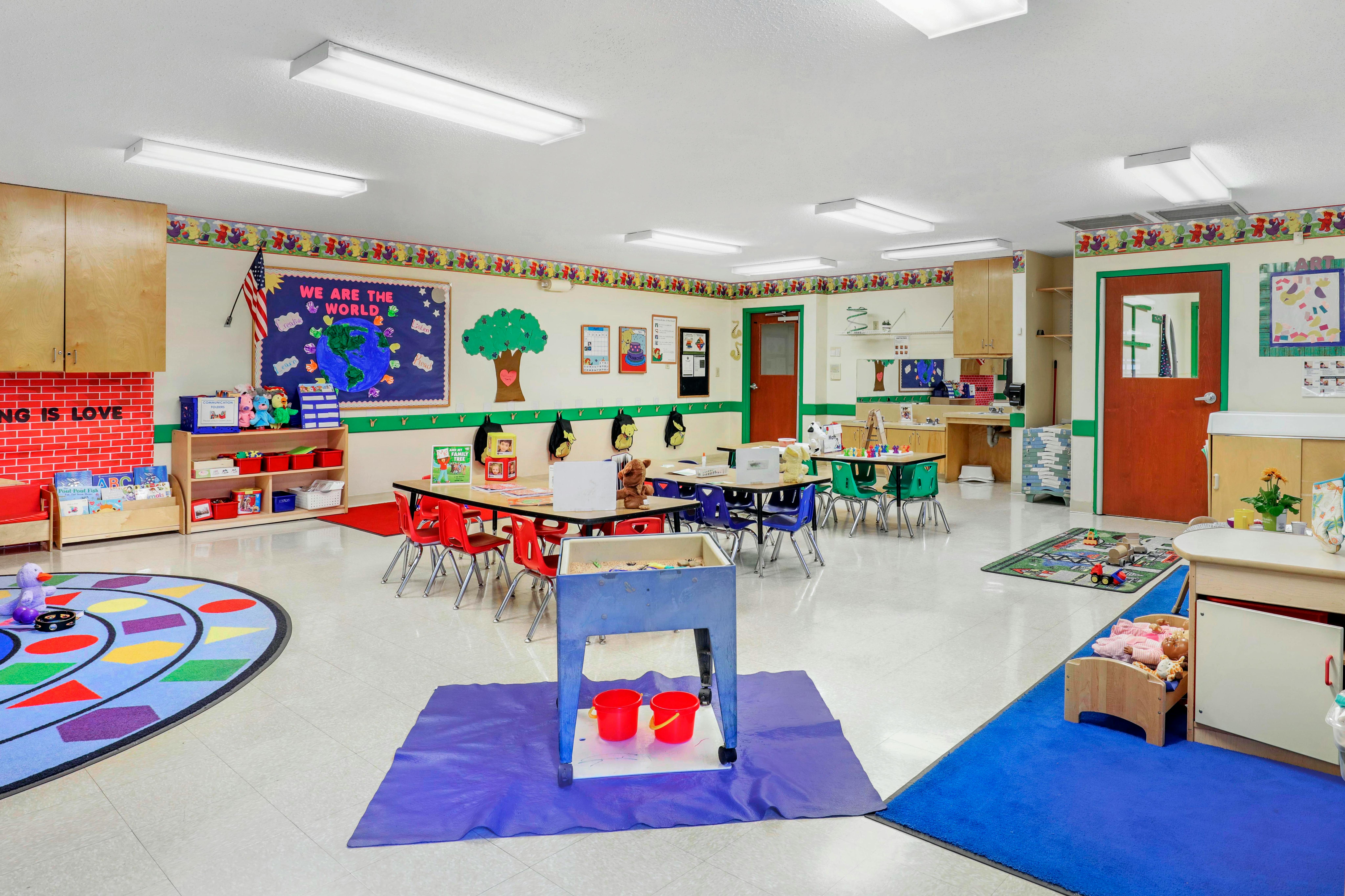 Classroom with child-sized tables and chairs, play mats, and a bulletin board that reads "We Are The World"