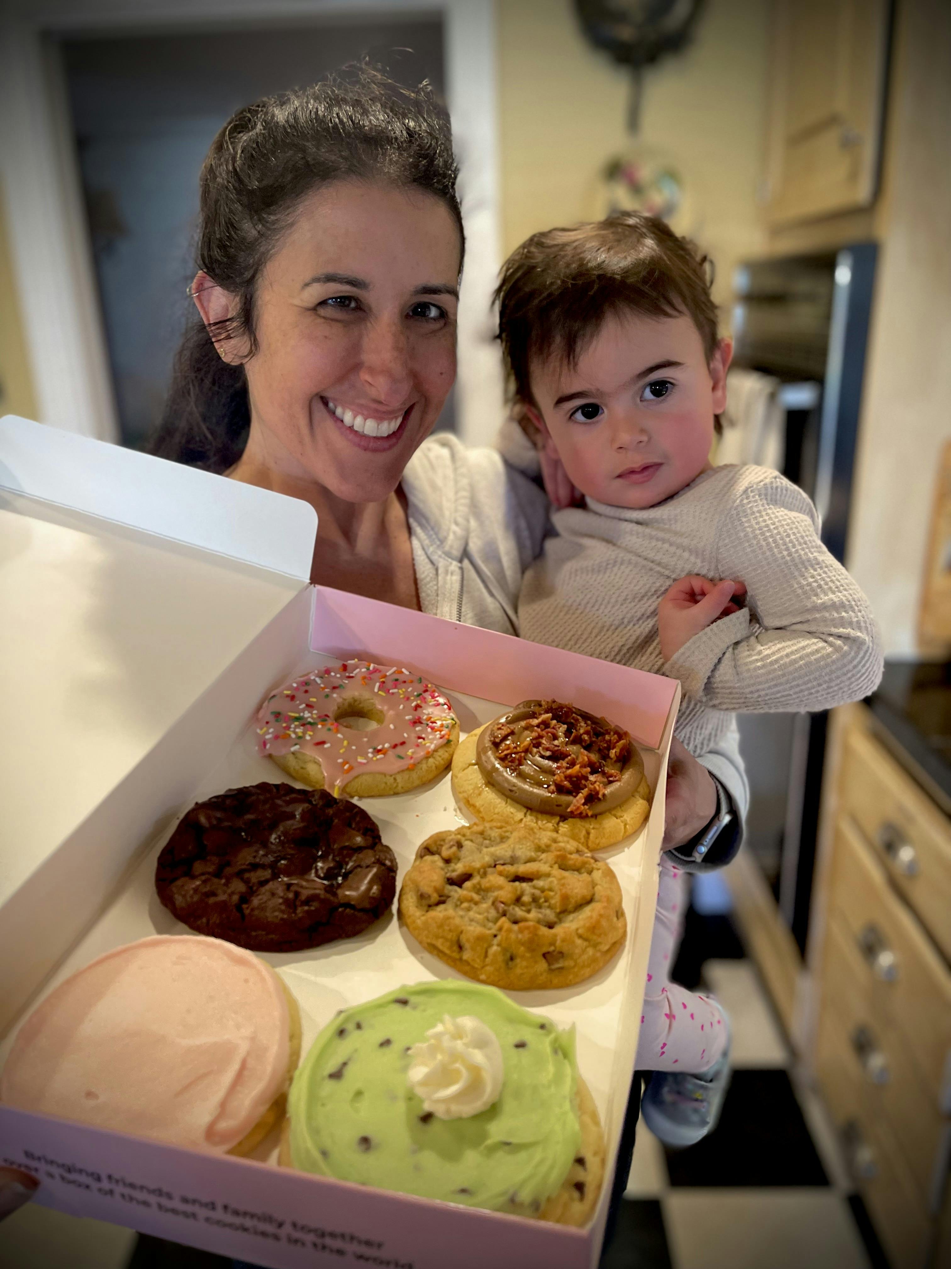 A person holding a child and a box of various frosted cookies
