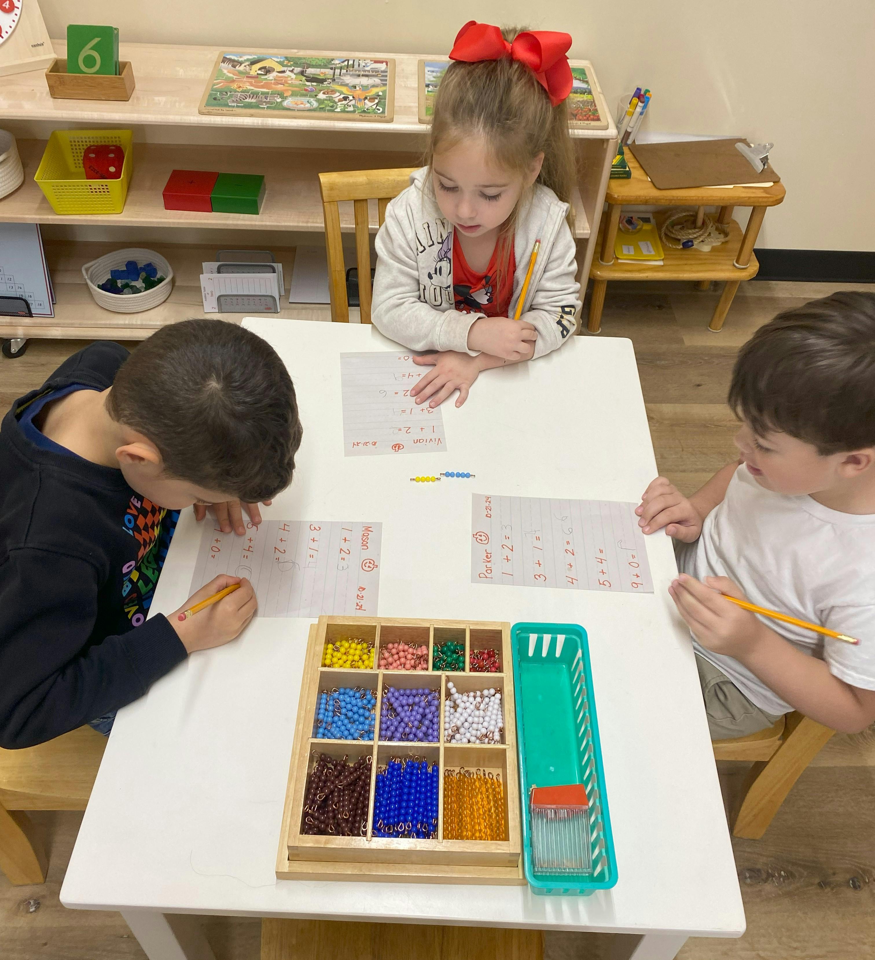 Three children writing at a table with a bead counting set