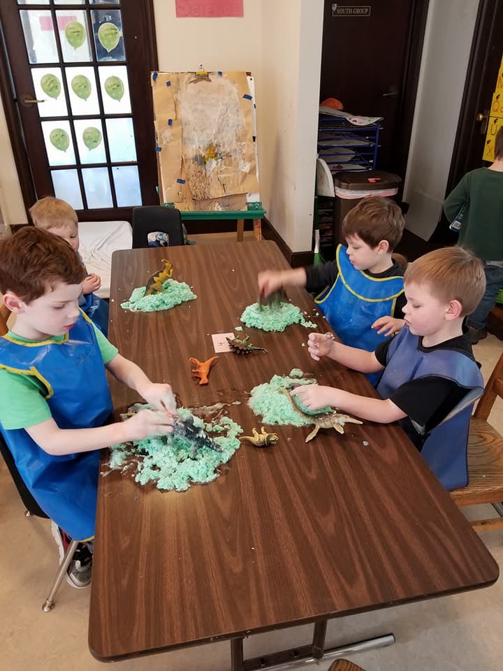 Children playing with green dough and toy dinosaurs at a table