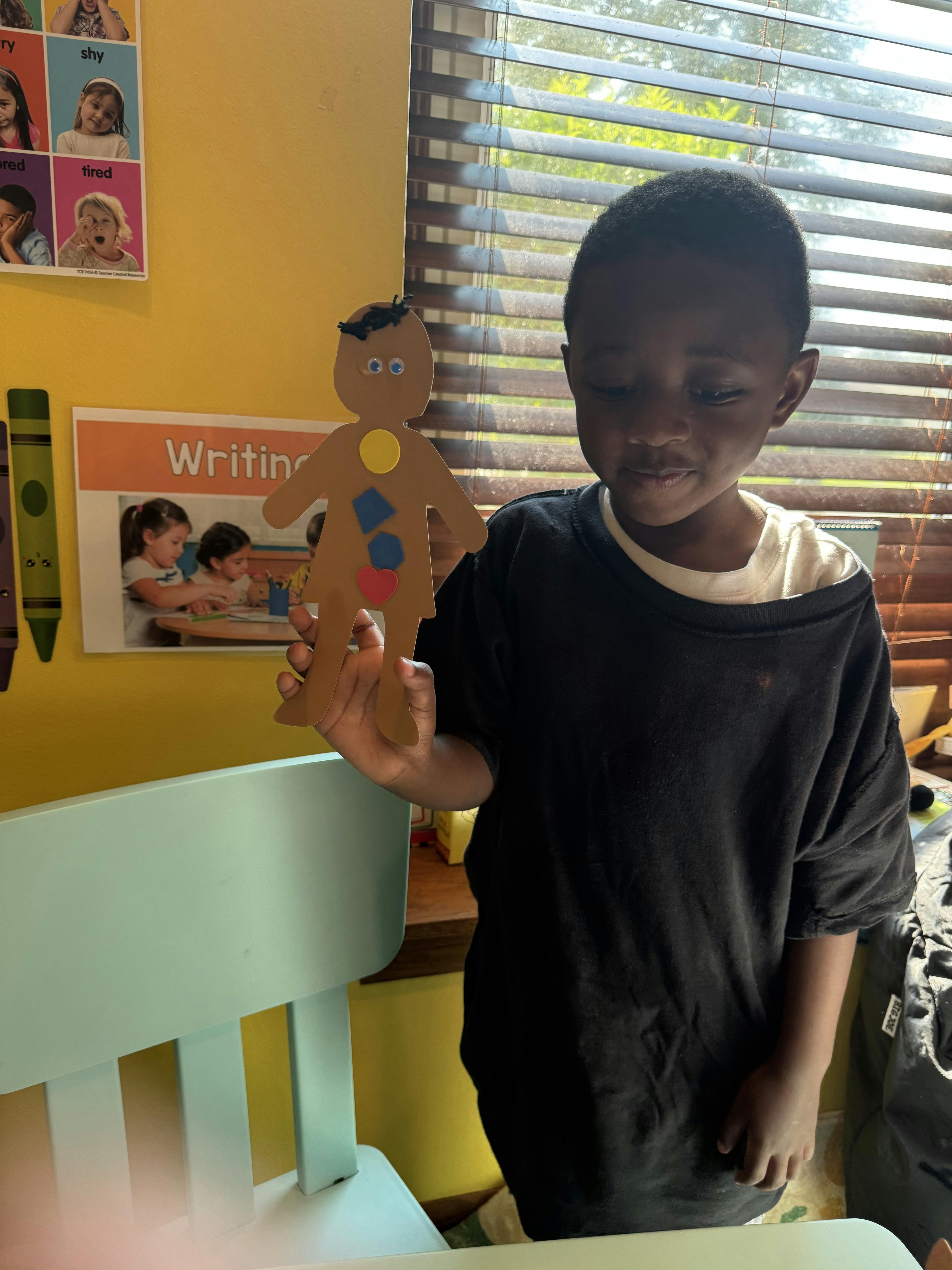 Child holding a paper cutout person with shapes in a classroom