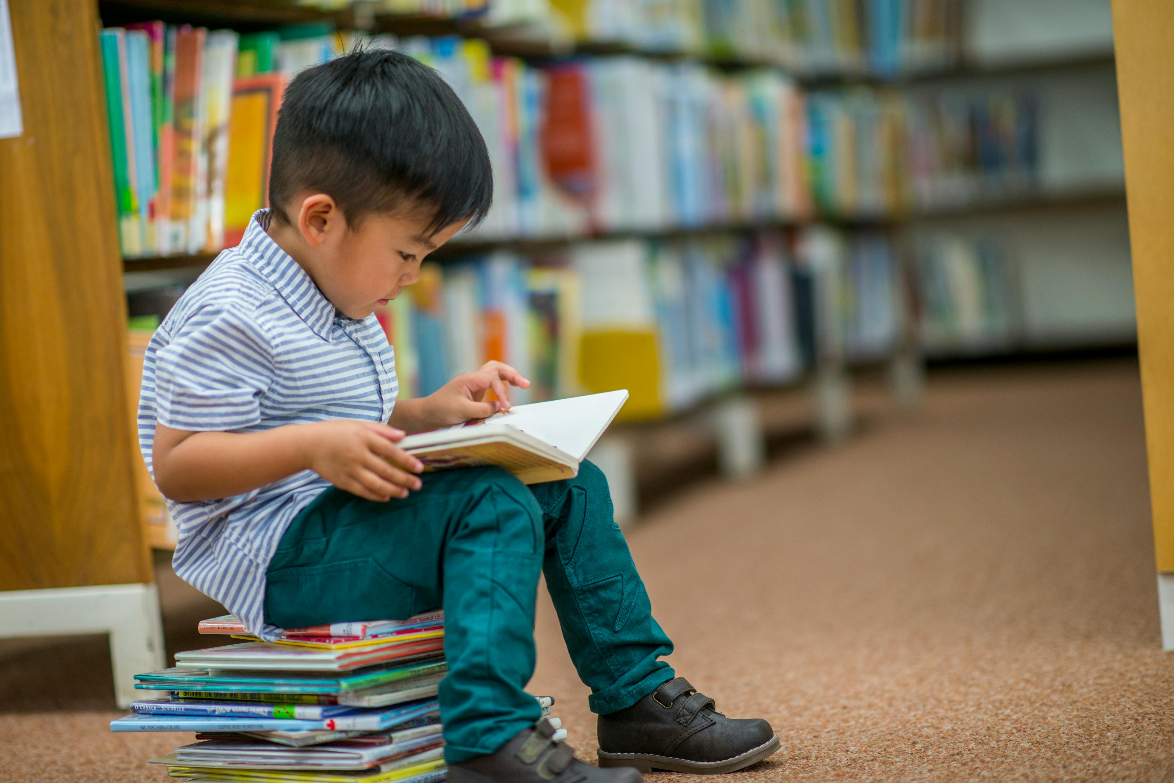 Child sitting on a stack of books reading an open book in a library