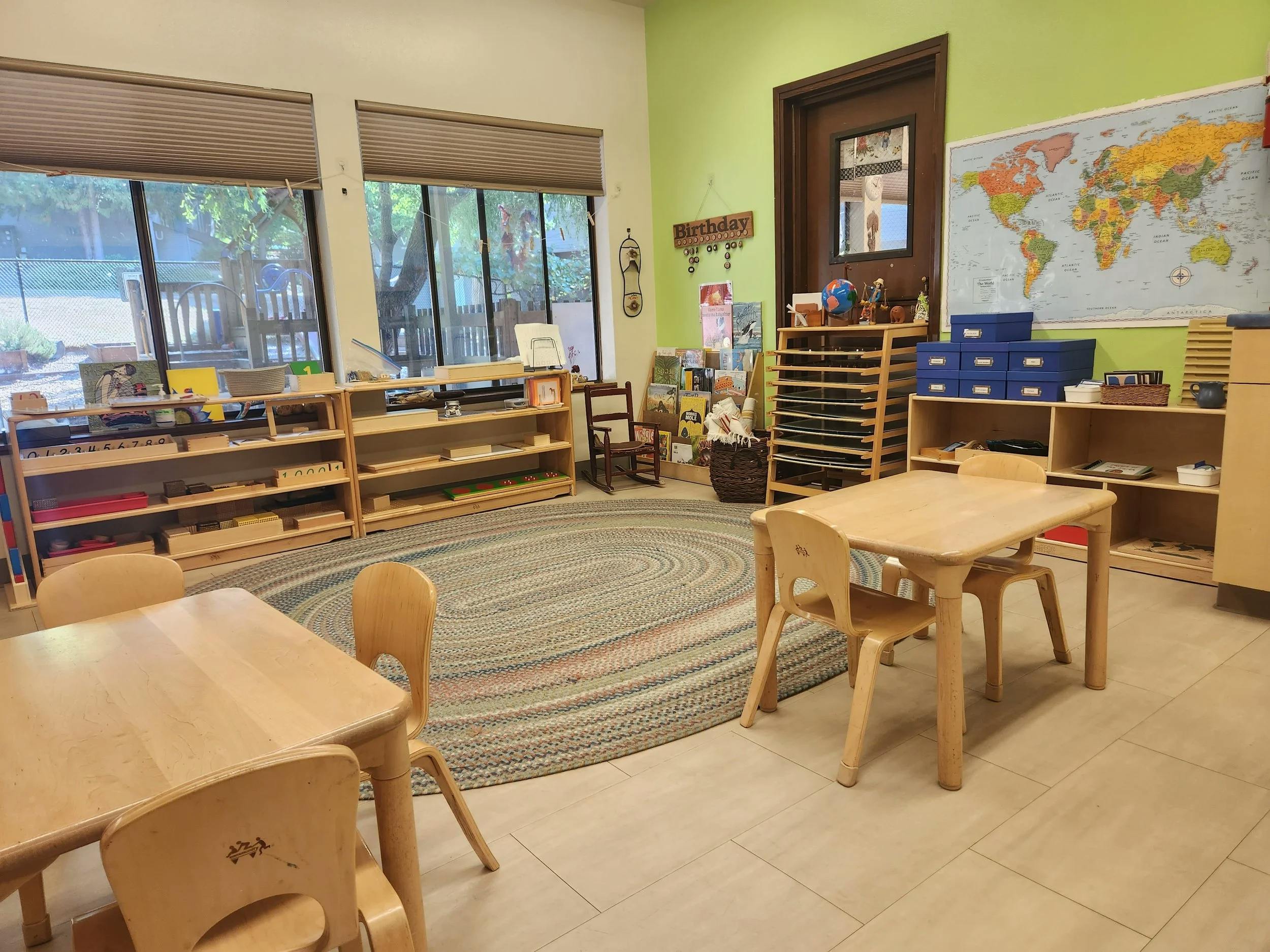 Classroom with small wooden tables and chairs, bookshelves, a world map, and windows
