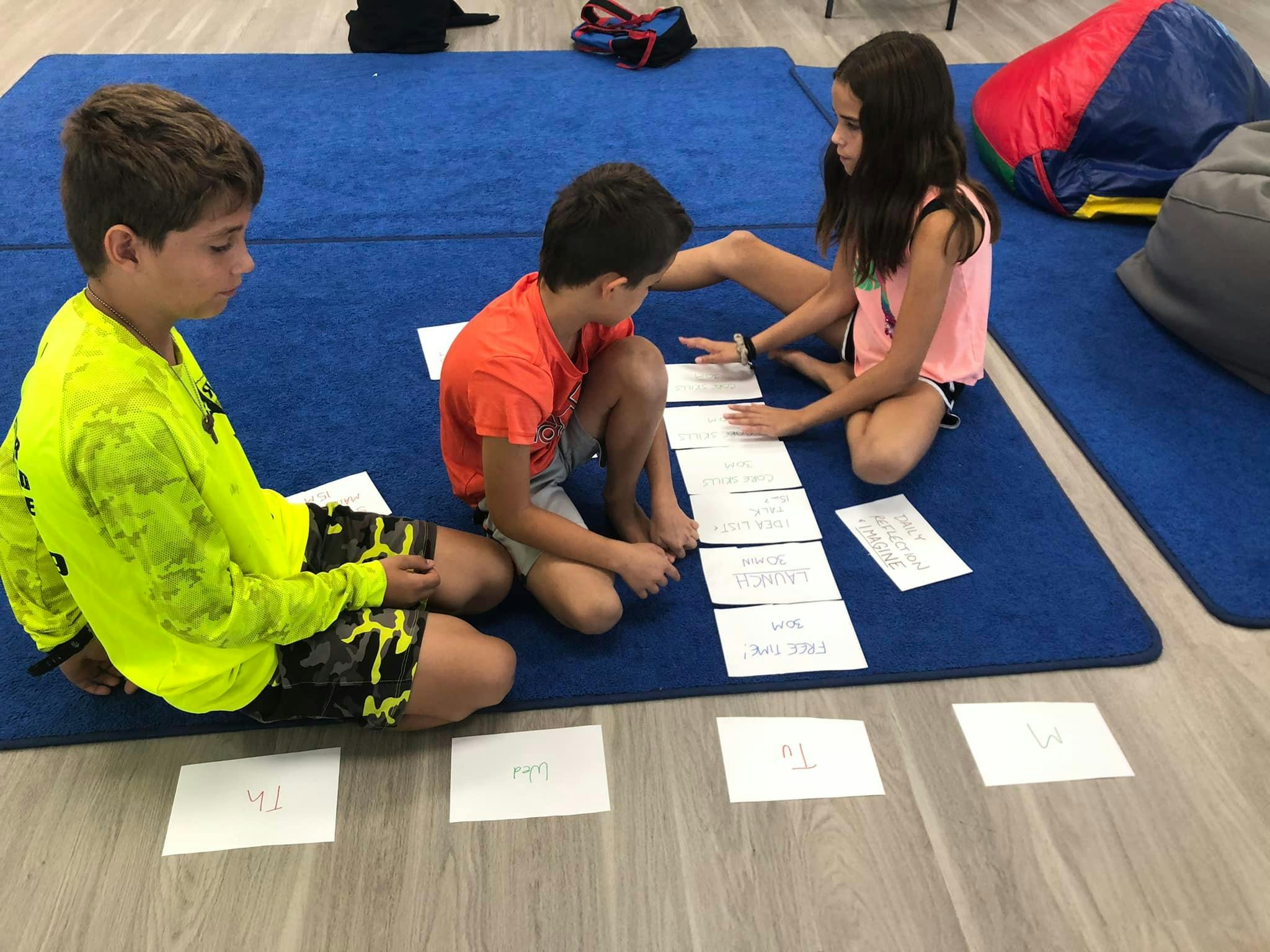 Three children sitting on a blue mat, looking at papers on the floor