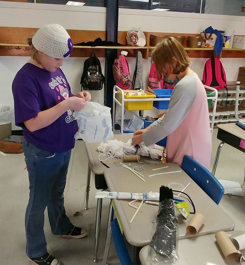 Two children working on a craft project at a desk in a classroom