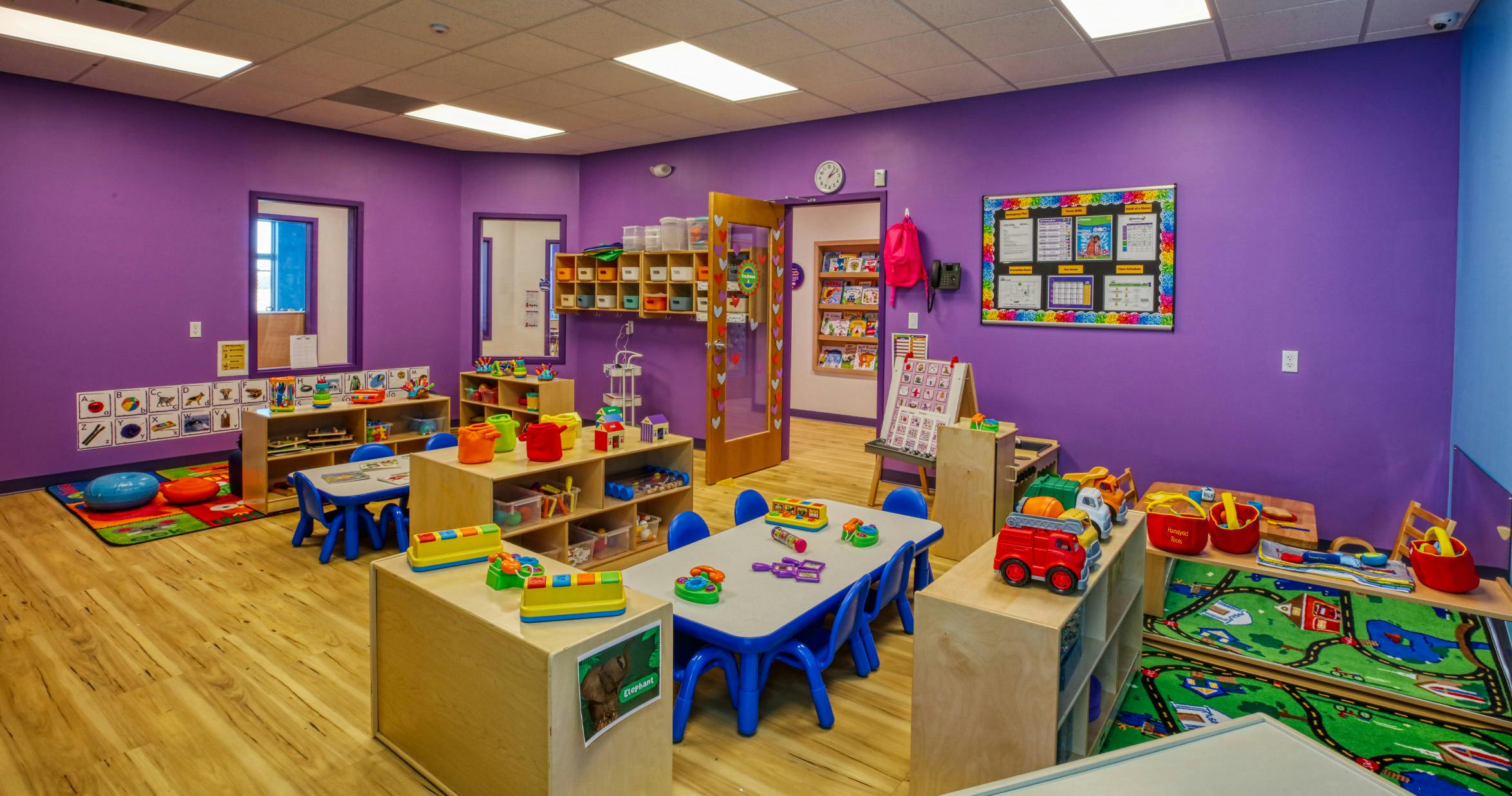 A brightly lit classroom with purple walls, tables, and toys
