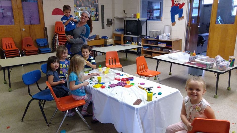 Adult holding a child, with other children at a table with colorful objects