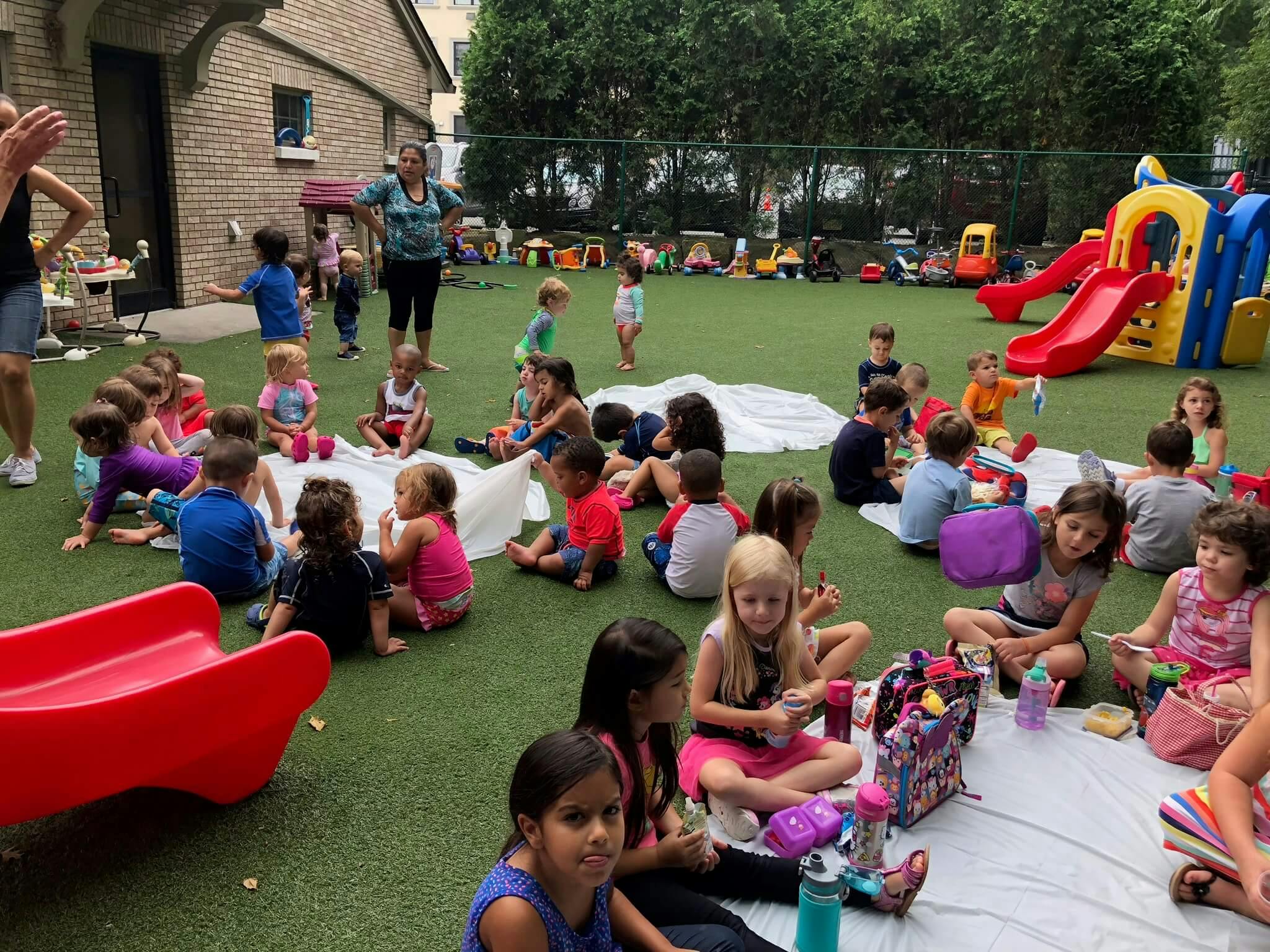 Many children and adults sitting on grass in an outdoor play area