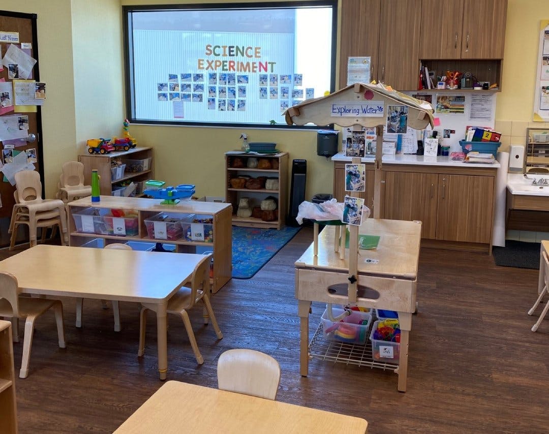 Classroom with small tables and chairs, toys, and "SCIENCE EXPERIMENT" on a window
