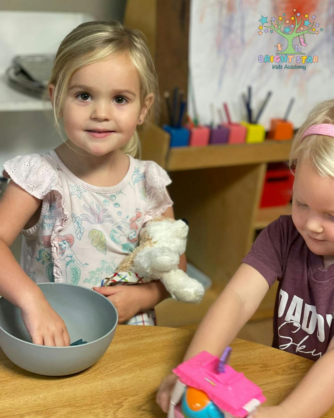 Child holding a teddy bear and reaching into a bowl, another child playing