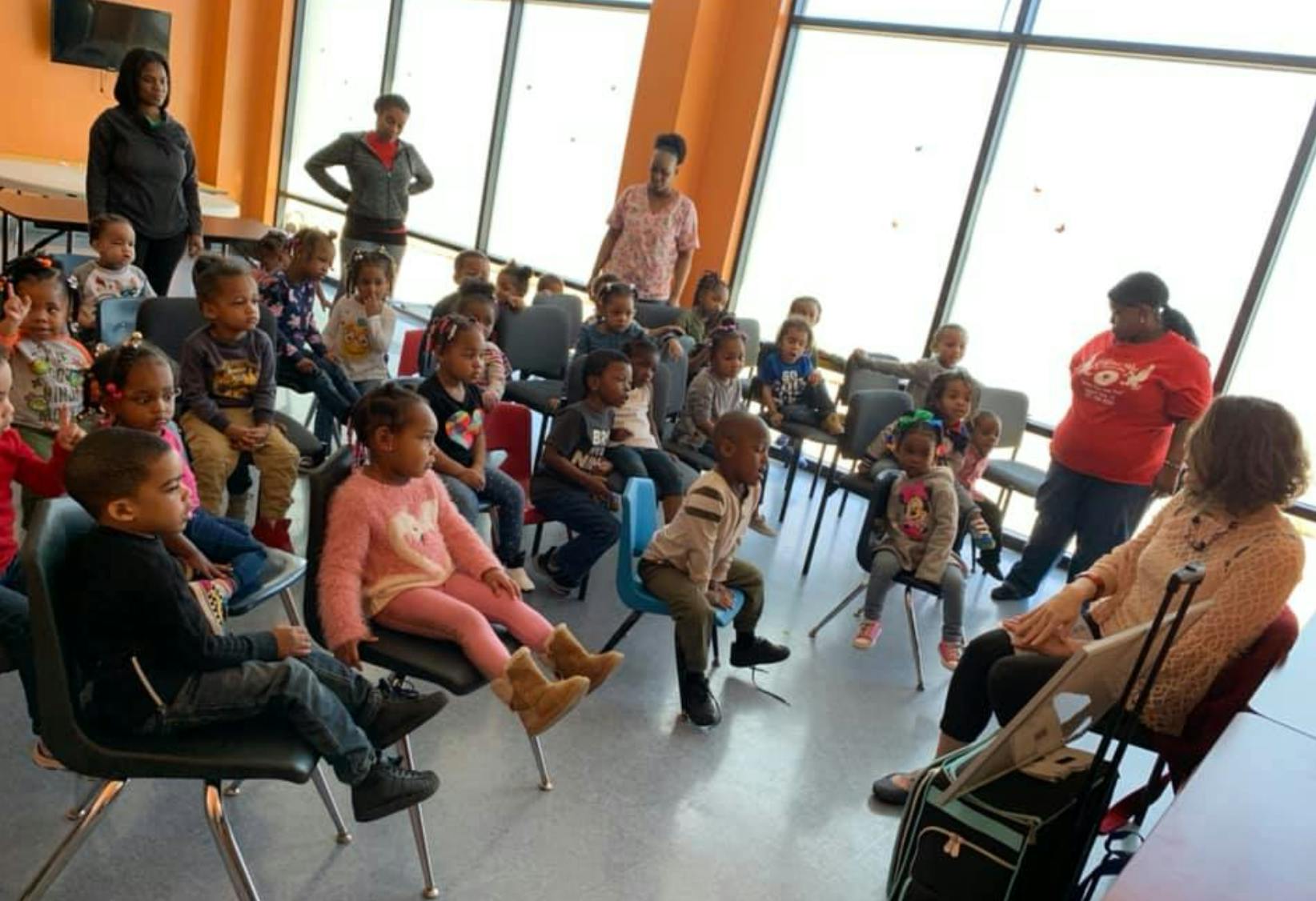 Children and adults sitting in chairs in a room with large windows