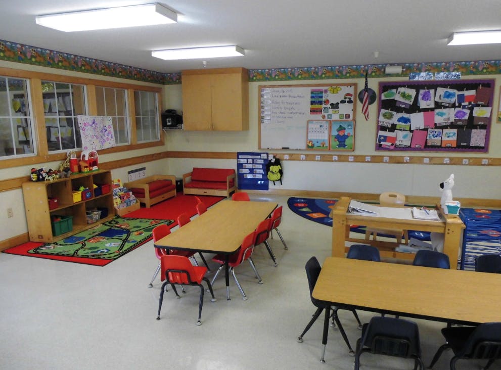 Classroom with small tables and chairs, play area with rug and toy shelf