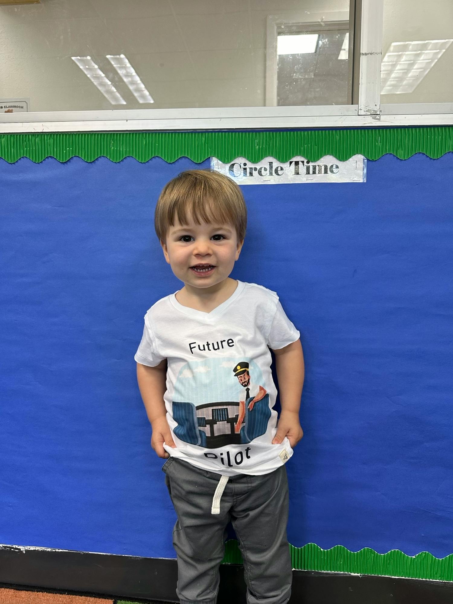 Child wearing a "Future Pilot" shirt standing in front of a blue wall with green wall